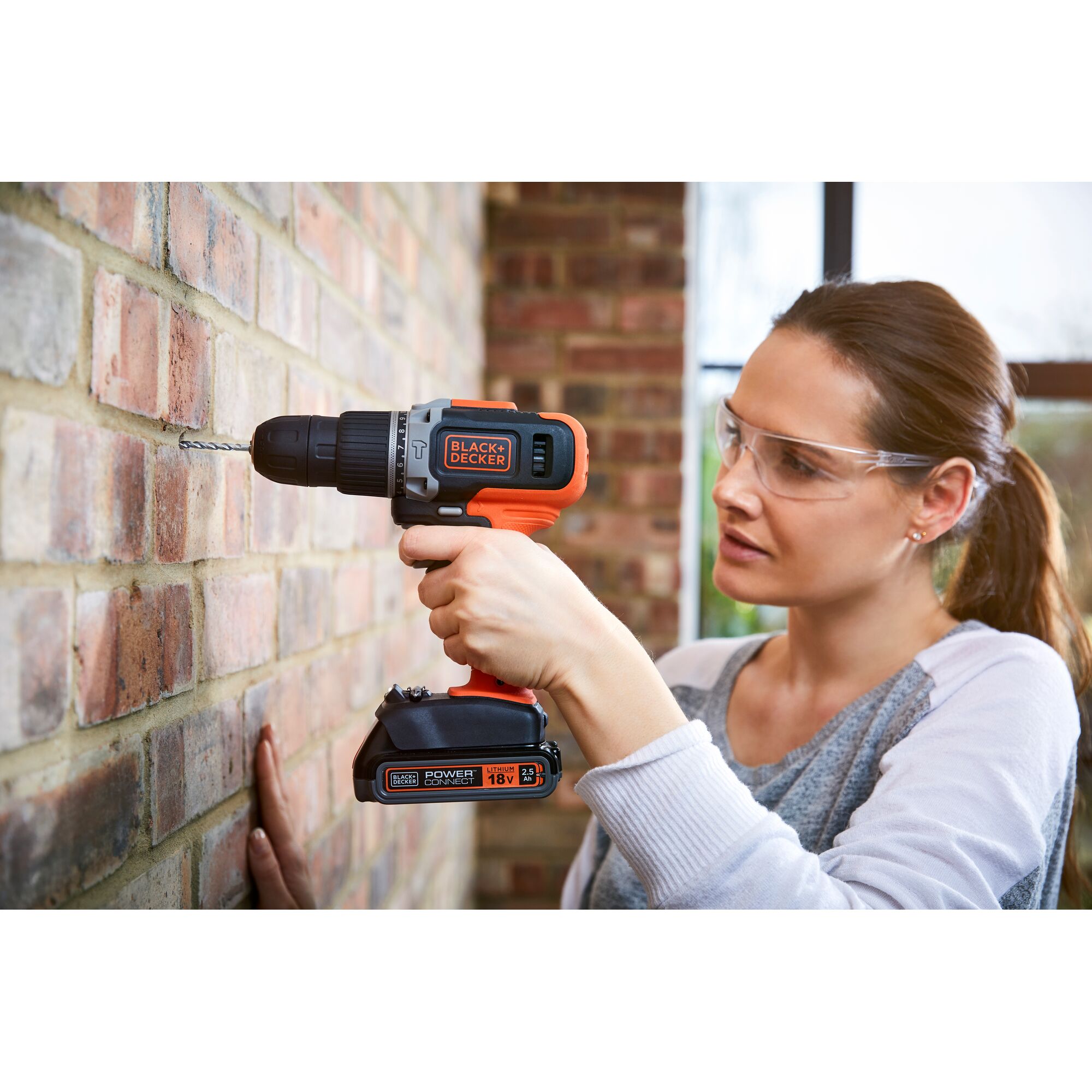 A person using a BLACK+DECKER cordless drill to drill into a brick wall. The individual’s face is blurred for privacy. The drill is orange and black, and the scene is set indoors.