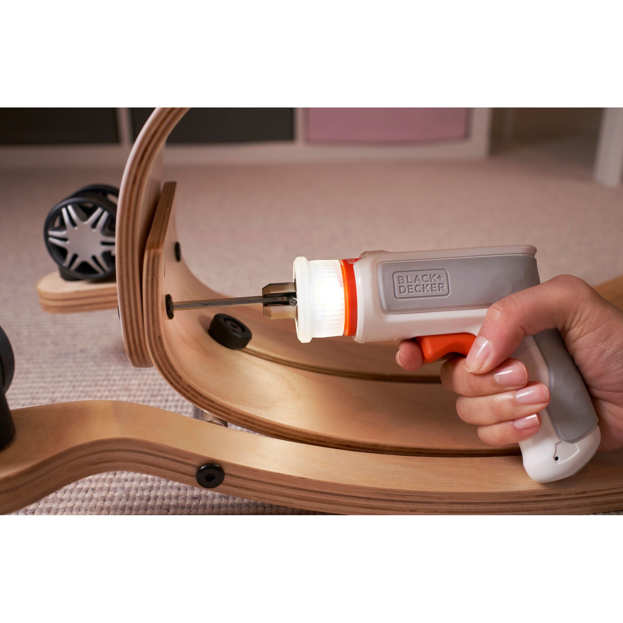 A close-up of a hand using a BLACK+DECKER electric screwdriver to assemble curved wooden pieces with screws. The screwdriver is white and gray with orange accents and is being used to fasten a screw into the wood.
