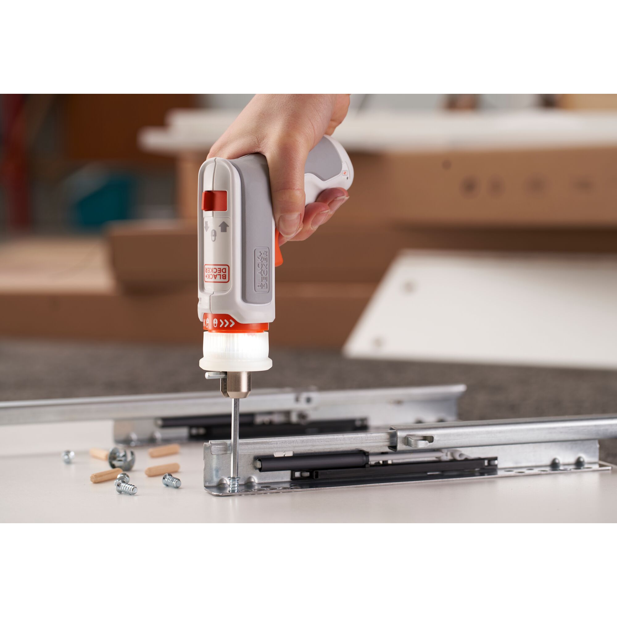 A hand using a BLACK+DECKER compact electric screwdriver to assemble metal furniture parts on a flat surface with screws and wooden dowels nearby.