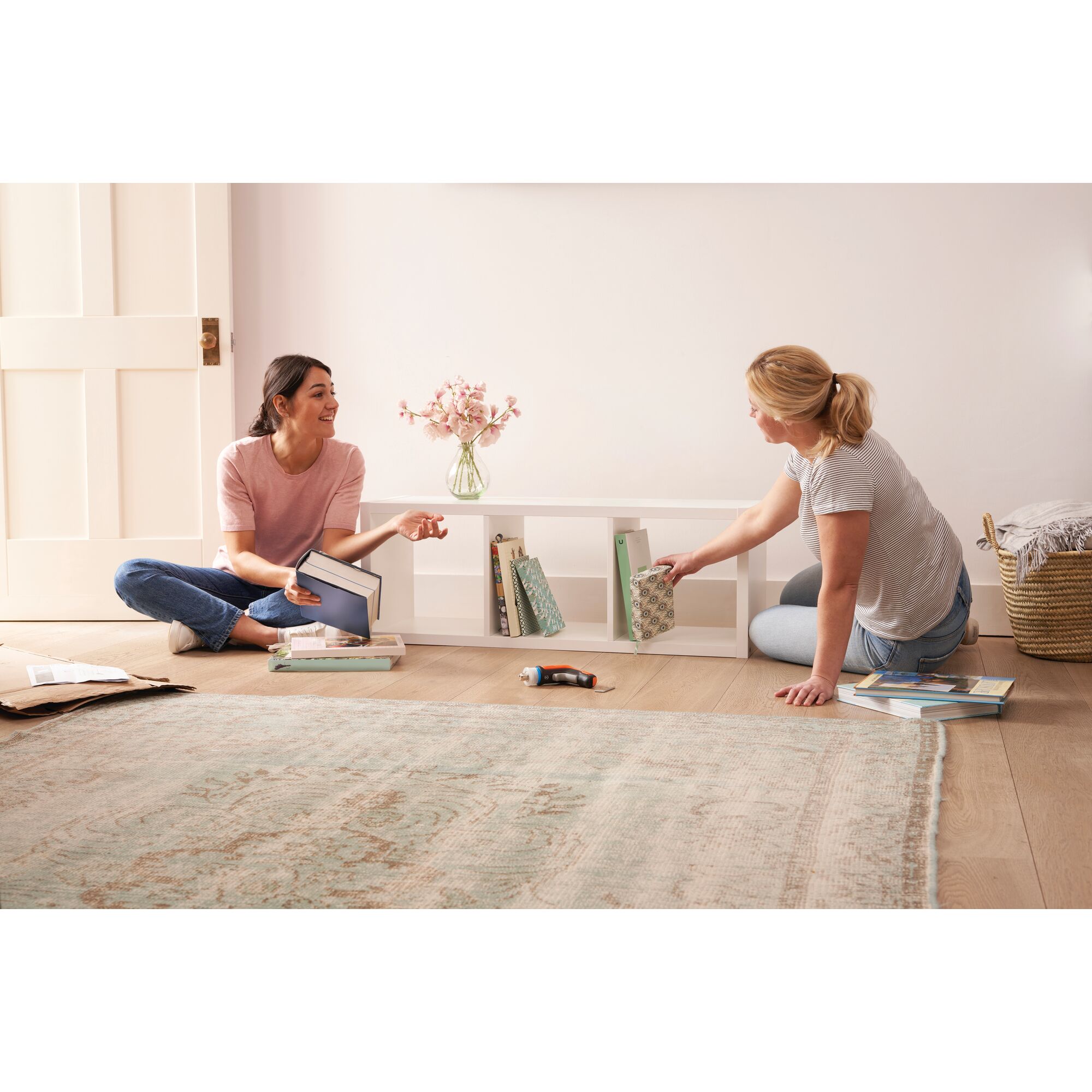 BLACKDECKER BCRTA601 cordless screwdriver on floor in living room, two women assembling white shelf with books.