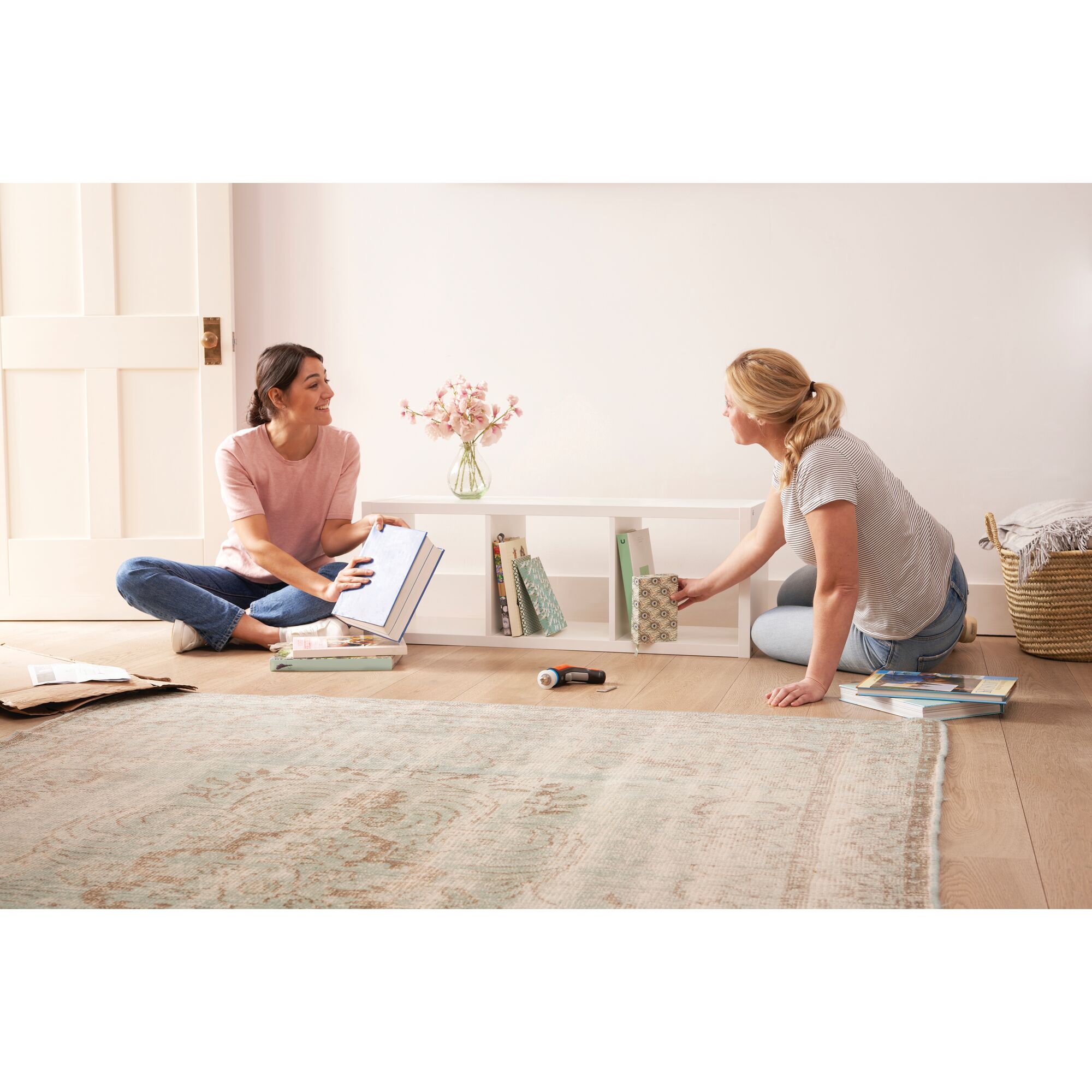 BLACKDECKER BCRTA601 cordless screwdriver on floor, two people organizing books on white shelf in bright room.