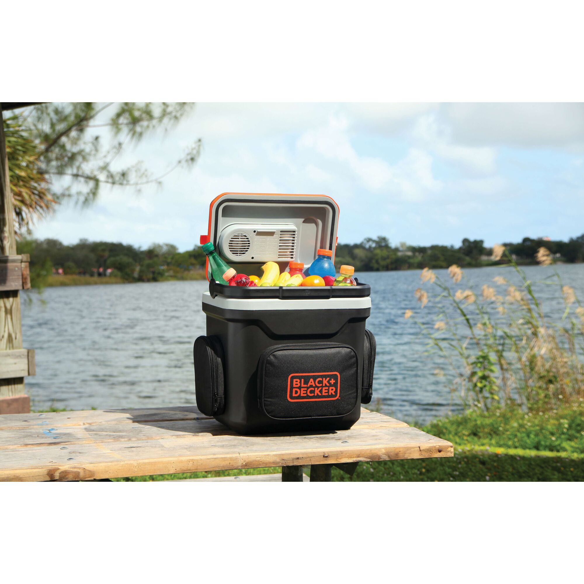 BLACKDECKER BDC24L portable cooler filled with drinks and fruit on a wooden table by a lake, outdoor setting.