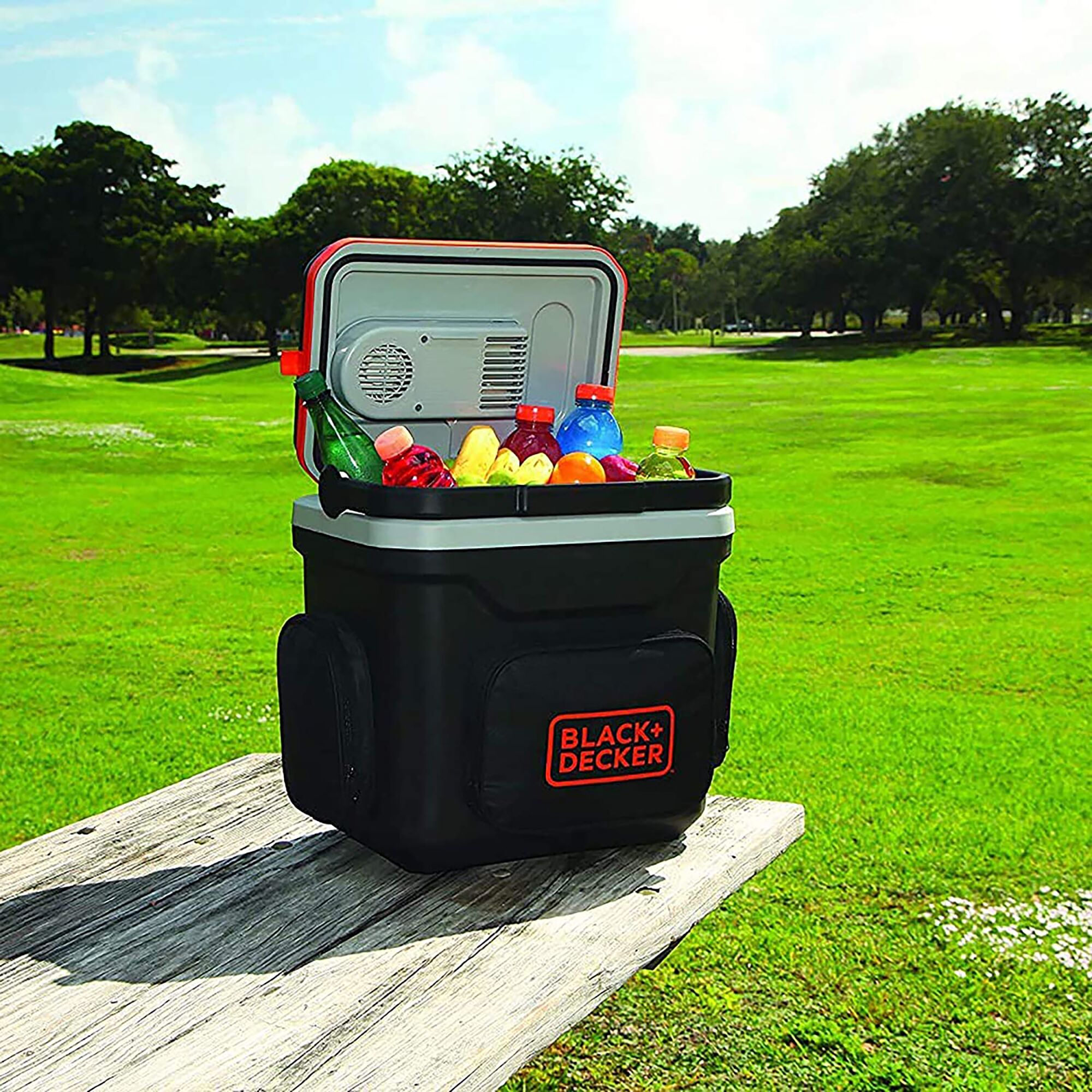 BLACKDECKER BDC24L portable cooler with open lid, filled with drinks and snacks, on park bench outdoors.