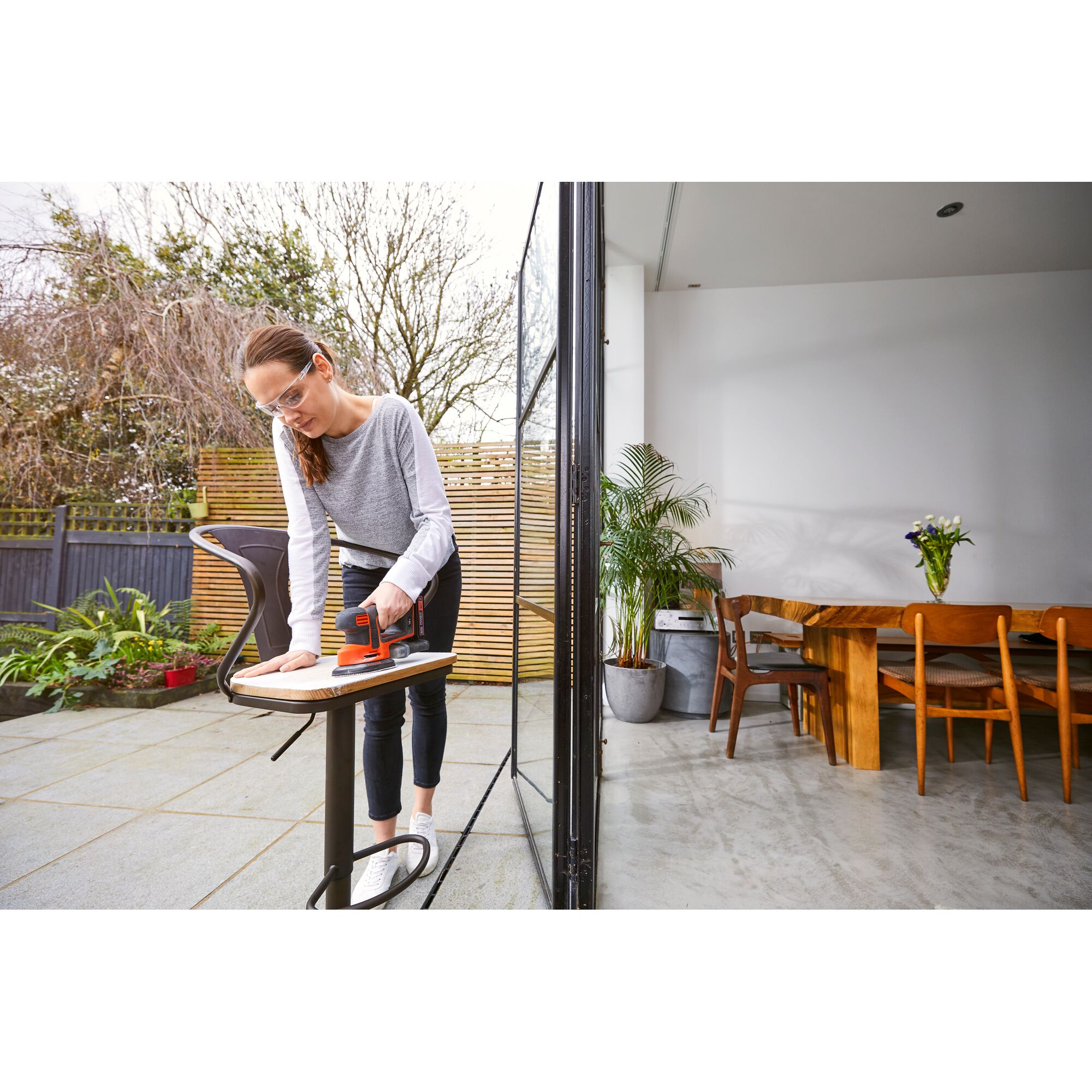 A person using a BLACK+DECKER BDCDS18 cordless sander on a piece of wood outside near a patio area. The scene includes modern outdoor and indoor furniture, plants, and a bright, airy home environment.