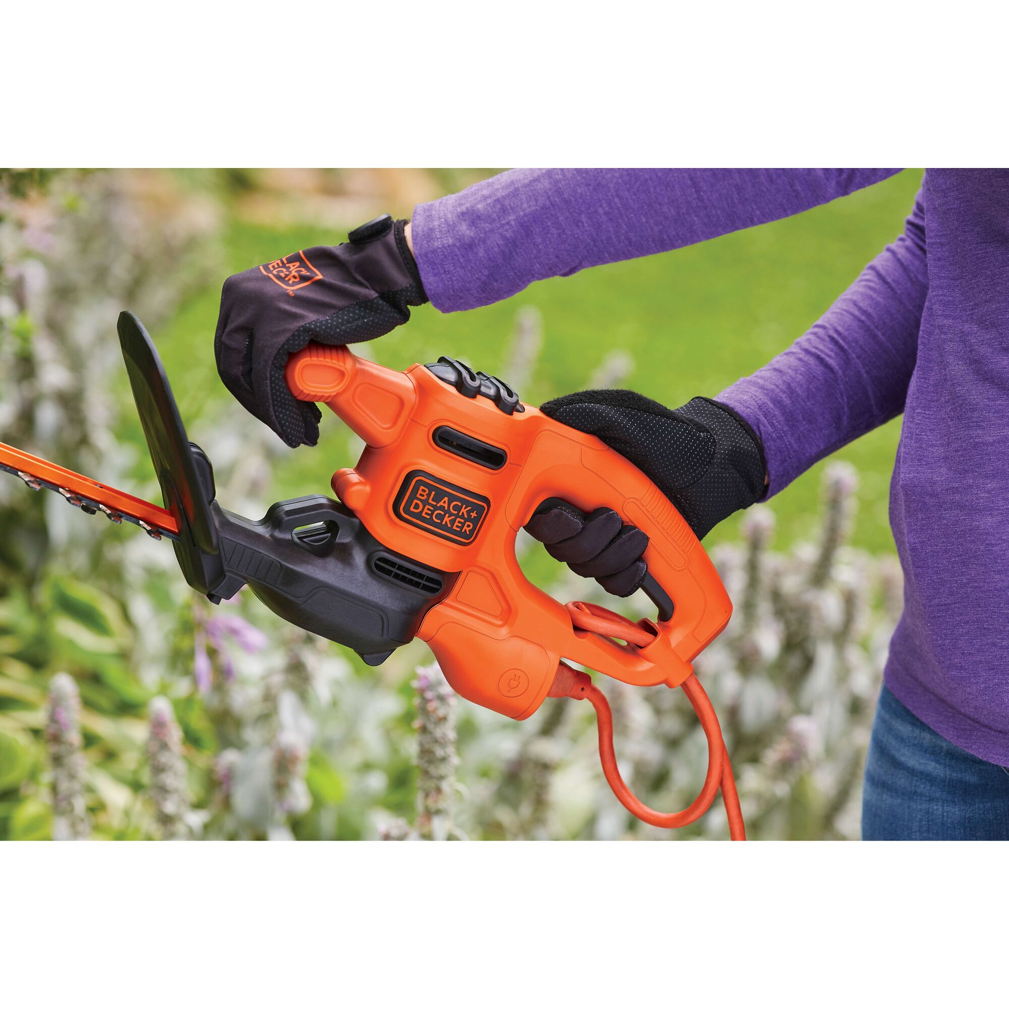 BLACKDECKER BEHT100 orange electric hedge trimmer held by gloved hands outdoors.