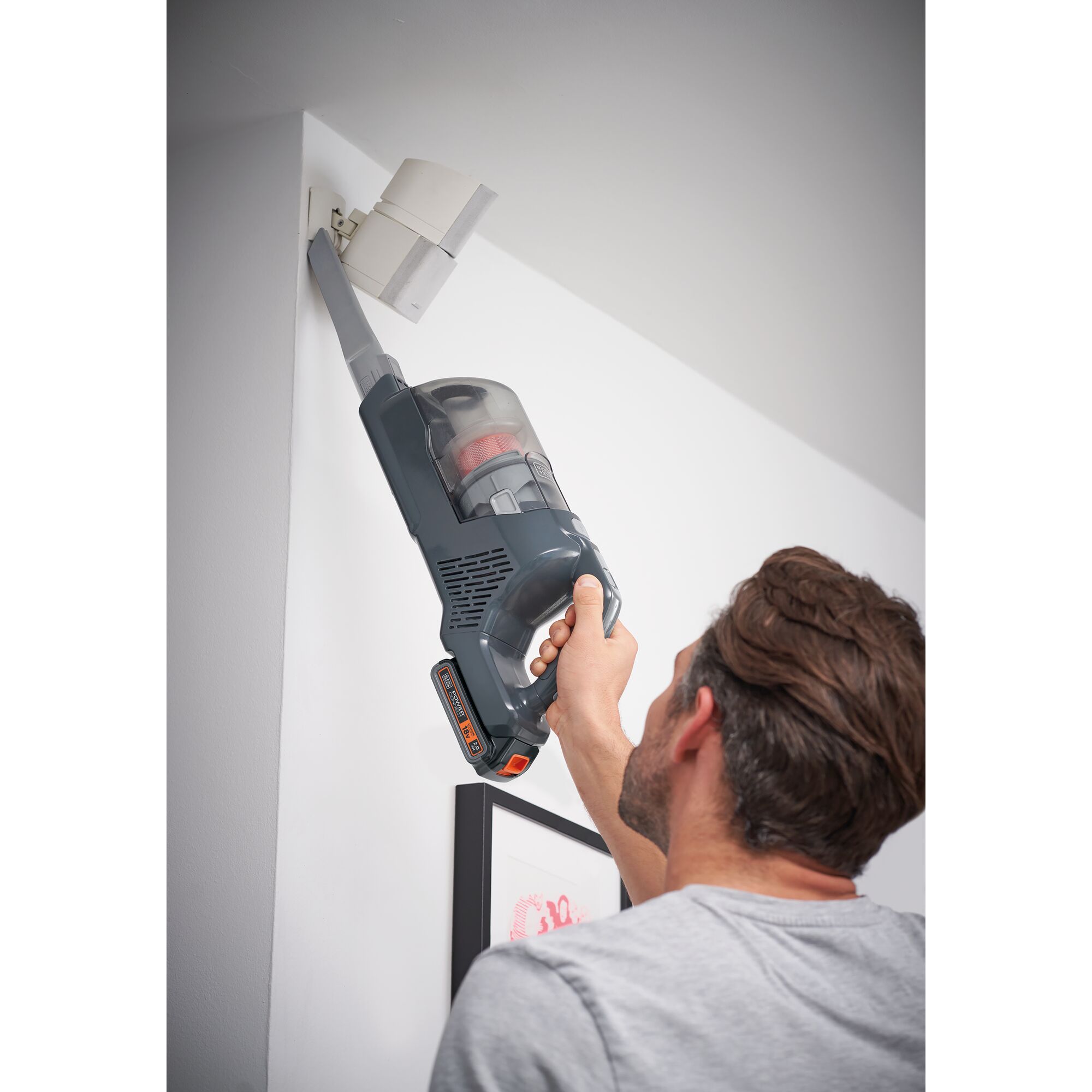 A person uses a BLACK+DECKER cordless vacuum cleaner with an extension nozzle to clean a high corner near the ceiling of a room.