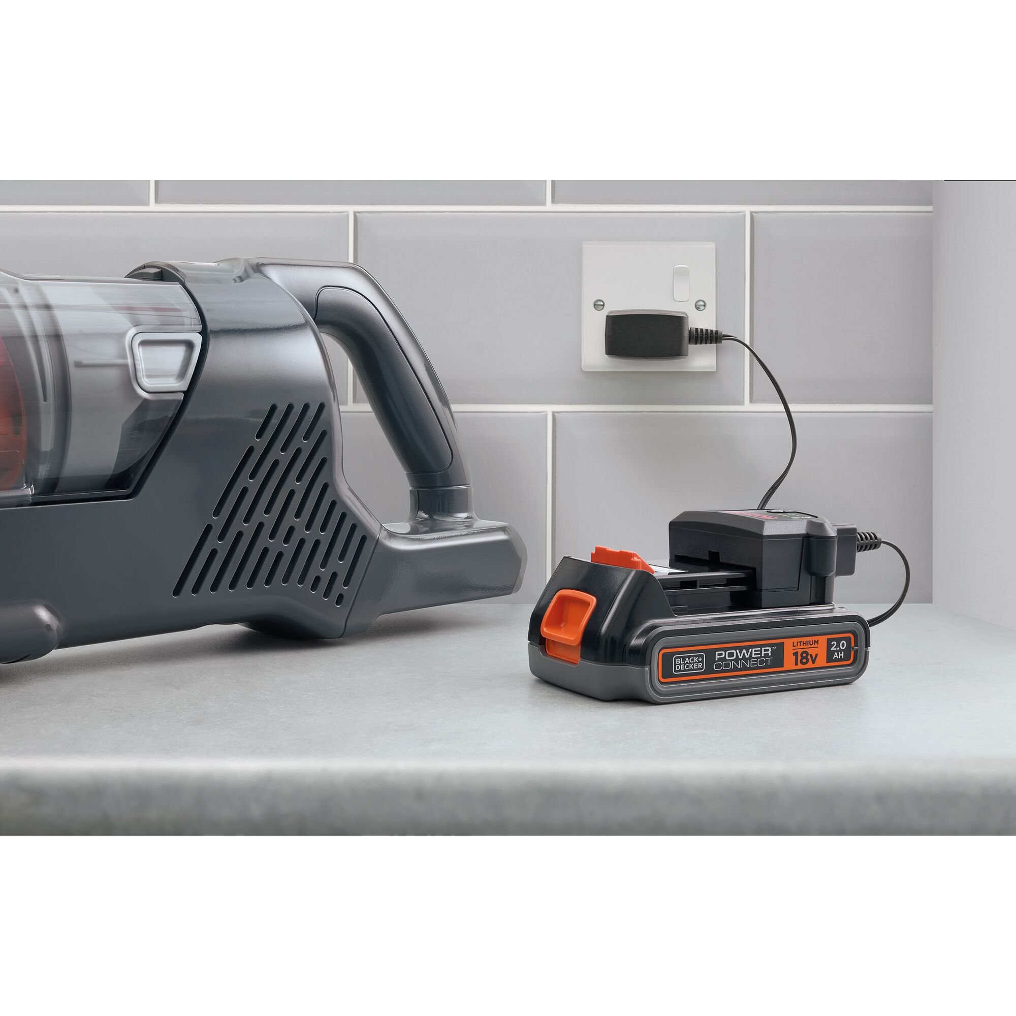 A BLACK+DECKER 18V lithium battery charger is placed on a countertop next to a cordless vacuum cleaner. The charger is plugged into a wall socket and is charging a battery. The scene is set against a tiled kitchen wall.