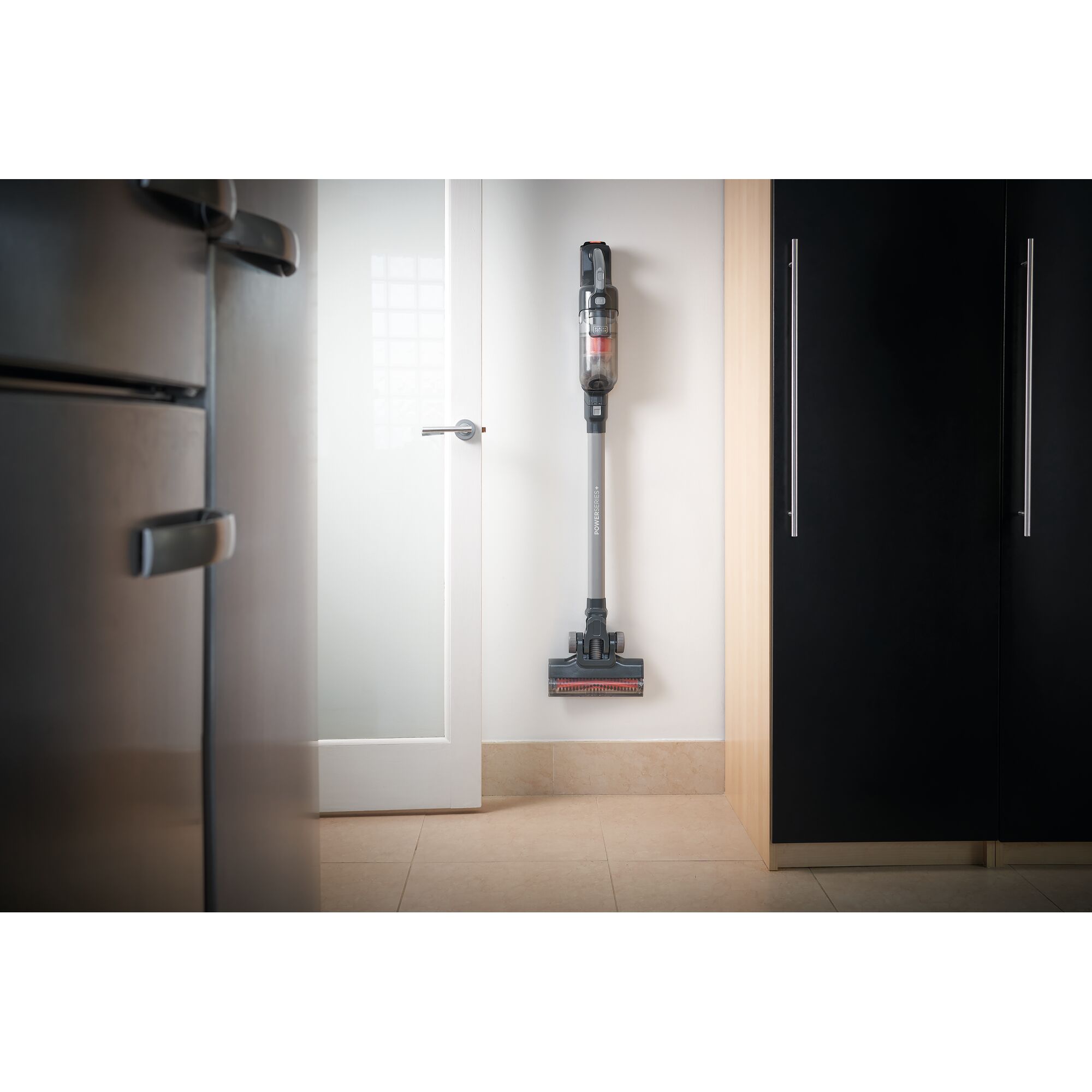 A cordless stick vacuum cleaner mounted on a wall in a modern kitchen, positioned between a refrigerator and a cabinet.