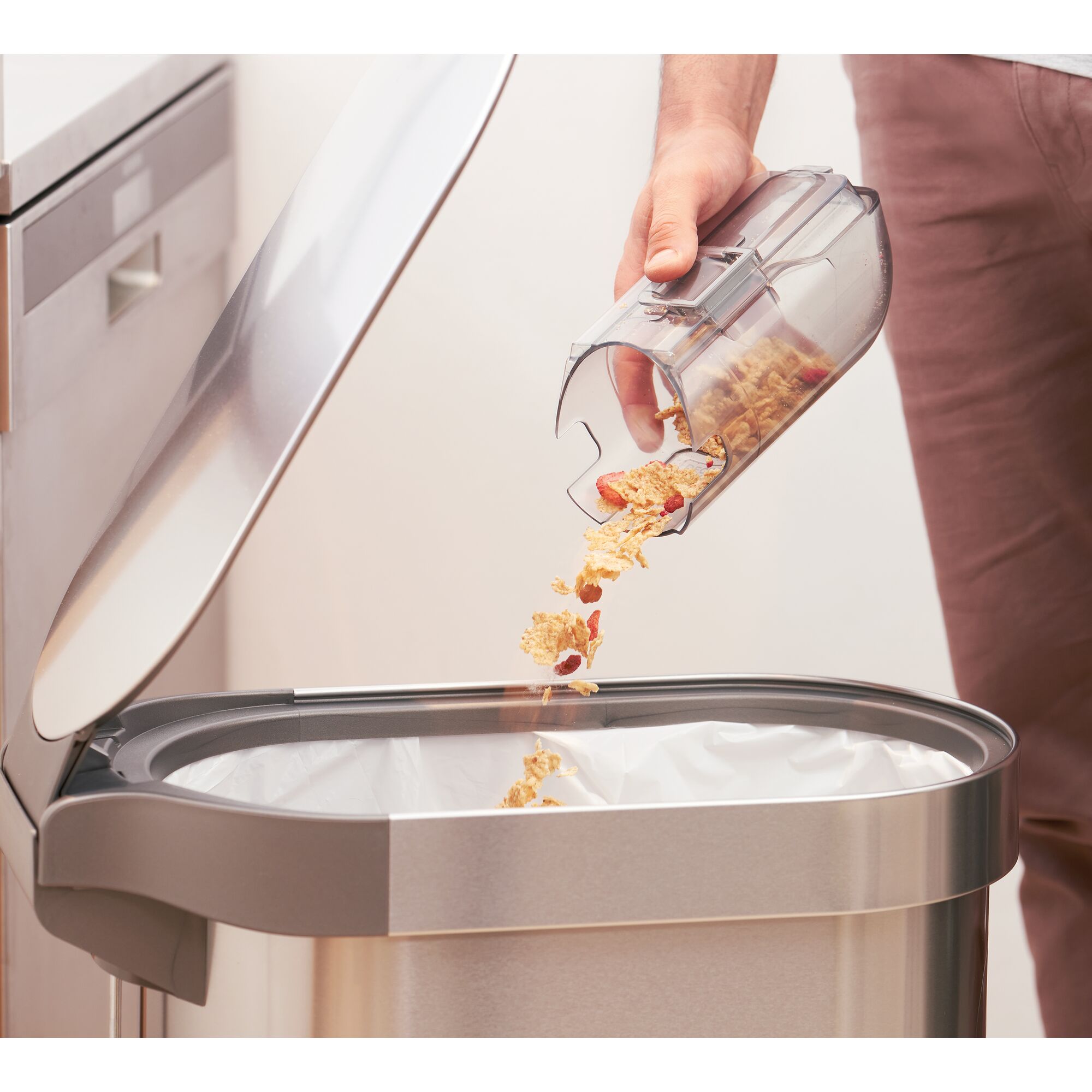 A person empties the contents of a vacuum cleaner dustbin, including cereal debris, into a stainless steel trash can with a lifted lid in a kitchen.