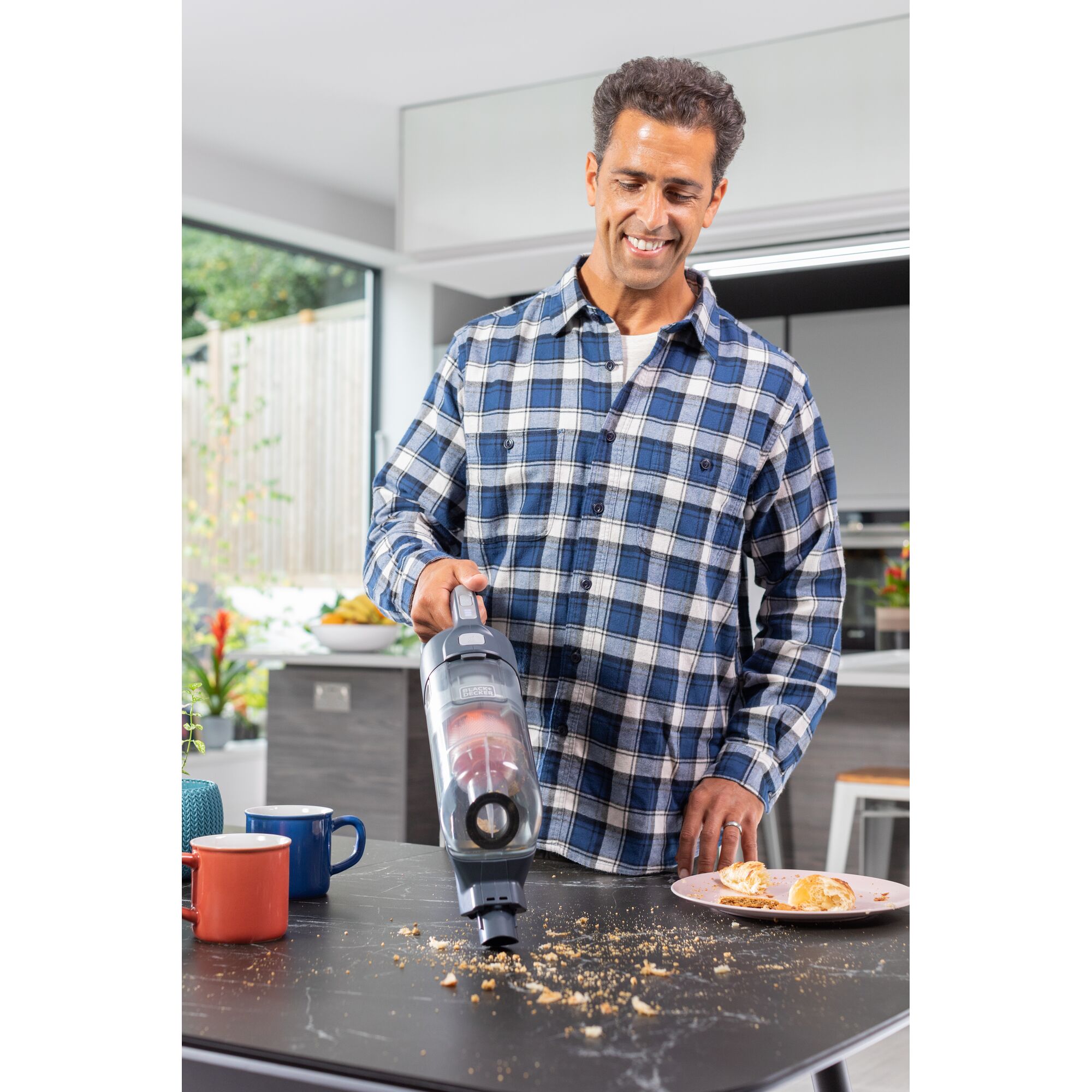 BLACKDECKER BHFEA520J handheld vacuum cleaning crumbs from kitchen table near mugs and pastry plate.