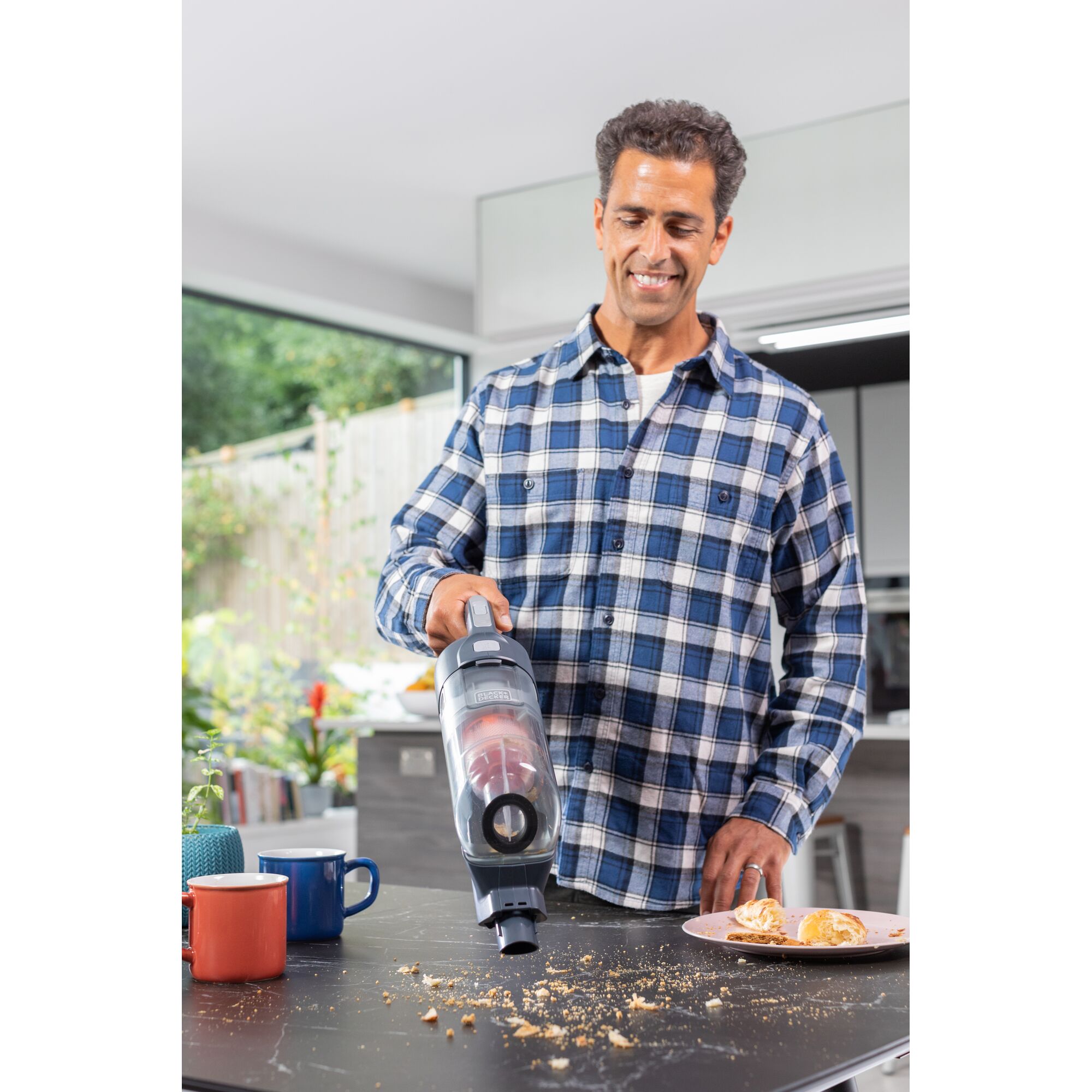 BLACKDECKER BHFEA520J vacuum cleaner removing crumbs from black kitchen table near mugs and pastry plate.