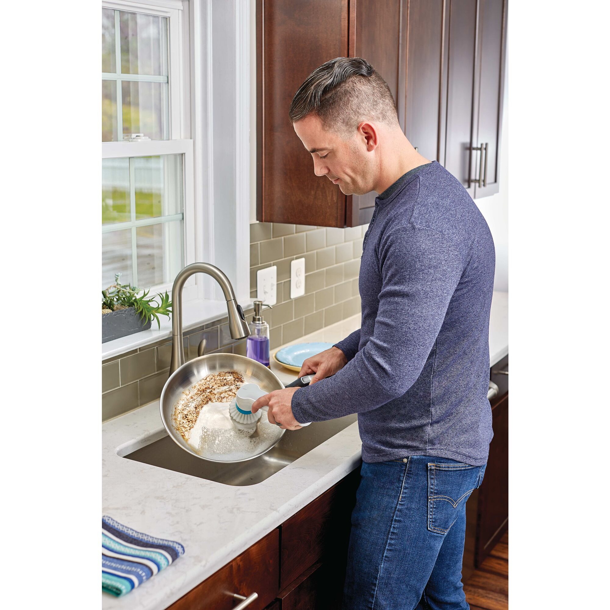 Grimebuster Pro rechargeable powered scrubber with charging stand being used by a person to wash a pan.