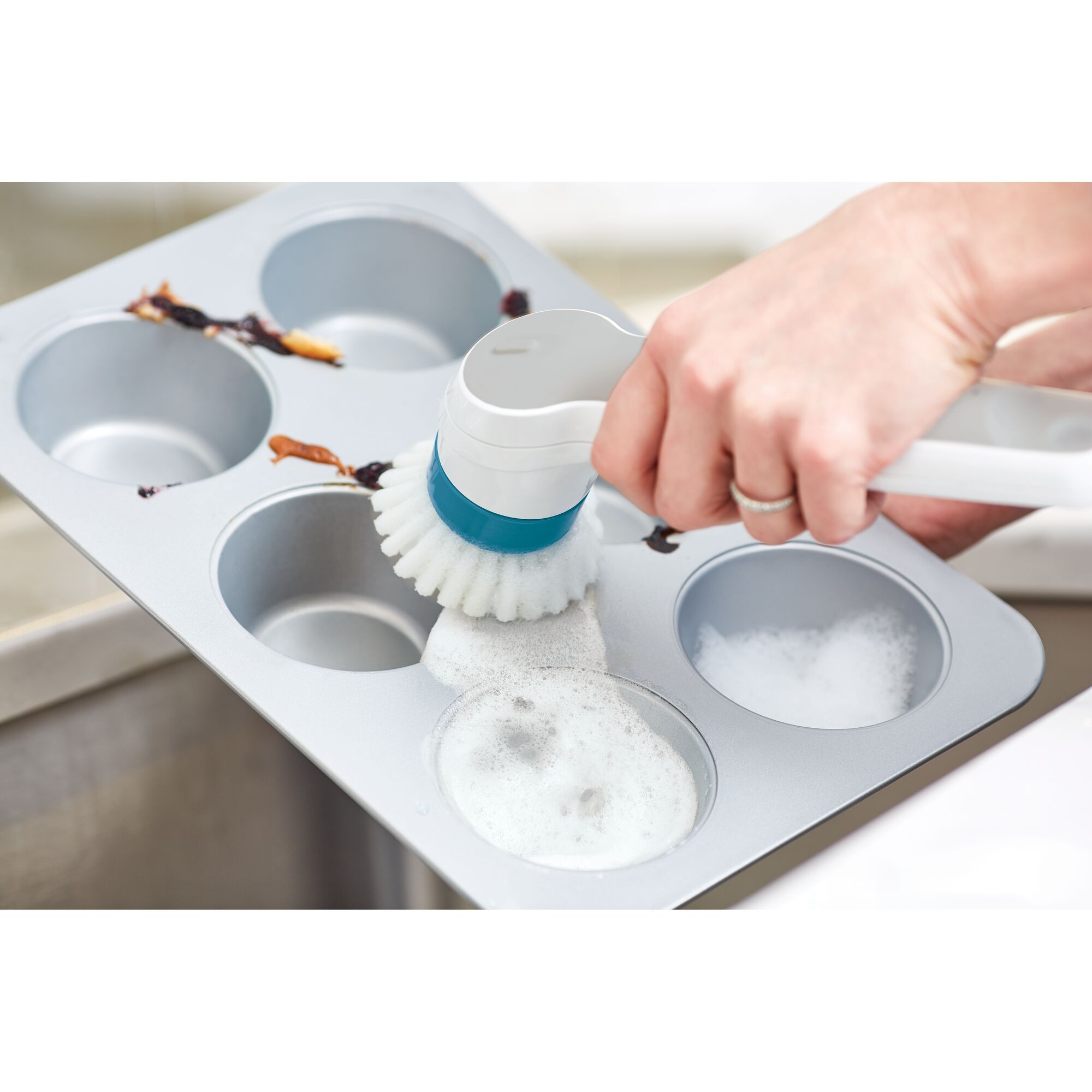 BLACKDECKER BHPC110 handheld brush cleaning muffin pan with soap suds, close-up angle.
