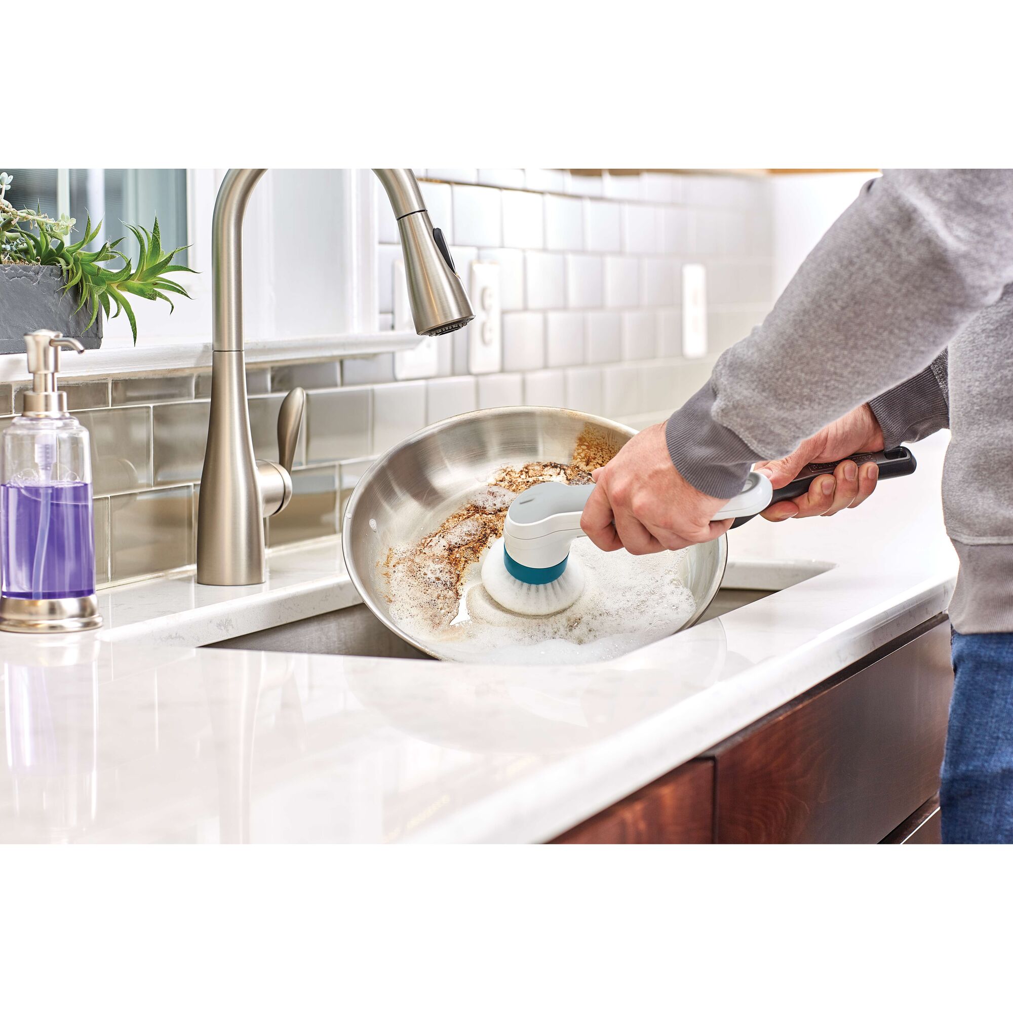 BLACKDECKER BHPC110 handheld scrub brush cleaning a stainless steel pan in kitchen sink.