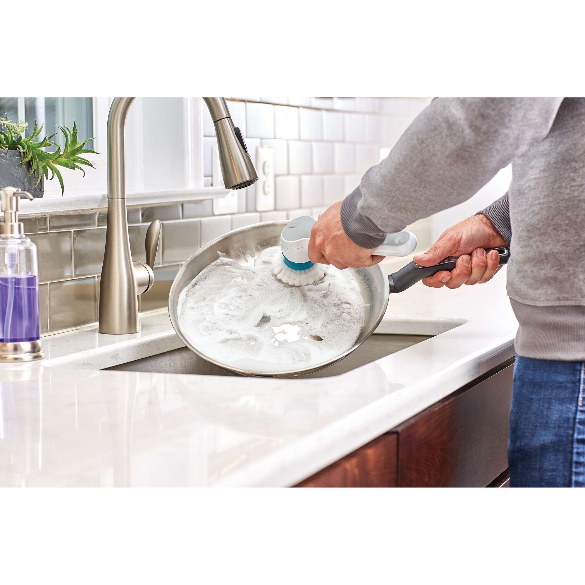 BLACKDECKER BHPC110 handheld scrub brush cleaning a soapy frying pan in a kitchen sink, side angle.