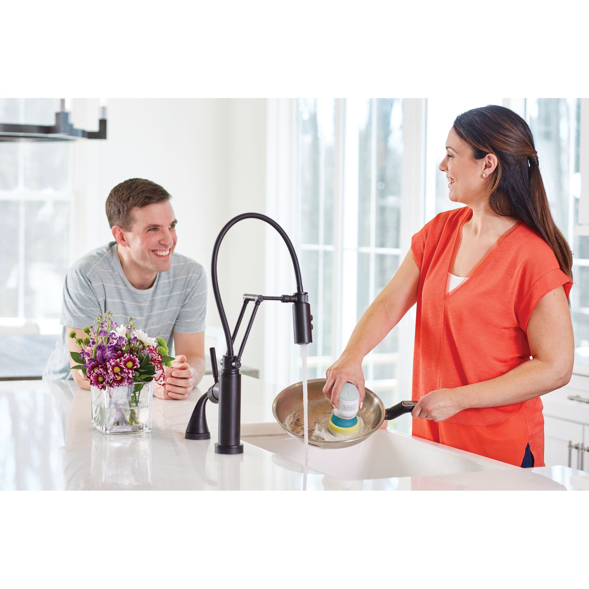 Photo of a woman using the BLACKDECKER BHPC130 scrubber to clean a pan in a kitchen sink, man sitting nearby.