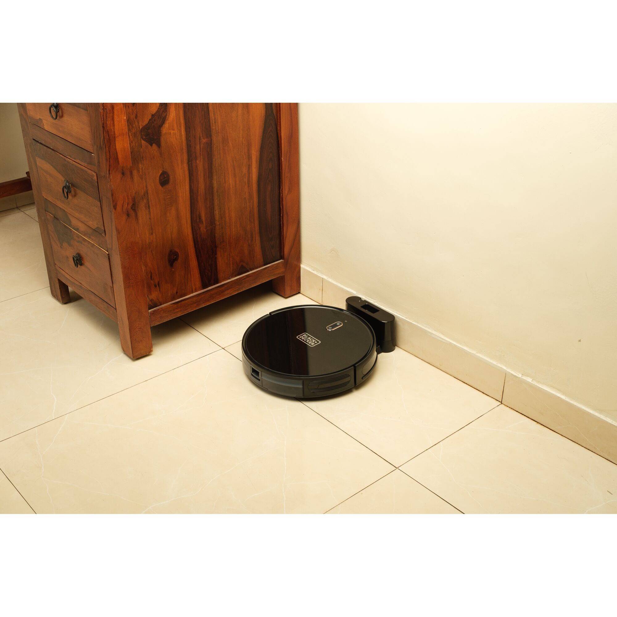 BLACKDECKER BRVA425B00 robot vacuum in black at charging dock next to wooden cabinet on beige tile floor.