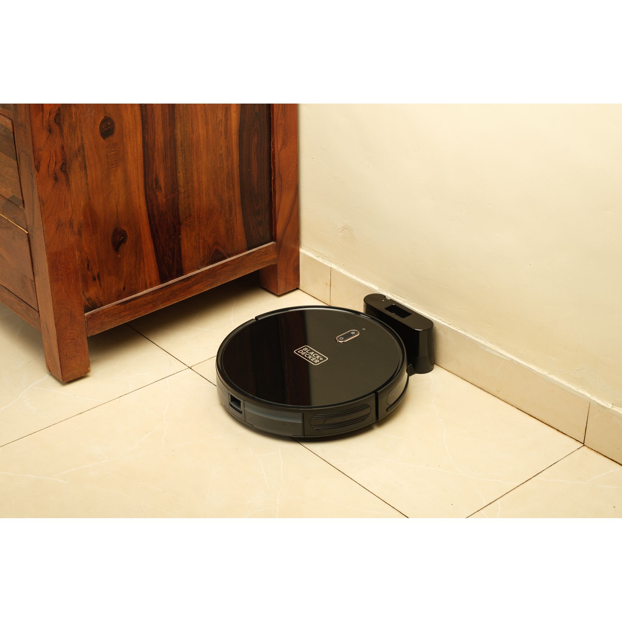 BLACKDECKER BRVA425B00 robot vacuum in black charging on dock next to wooden cabinet on tile floor