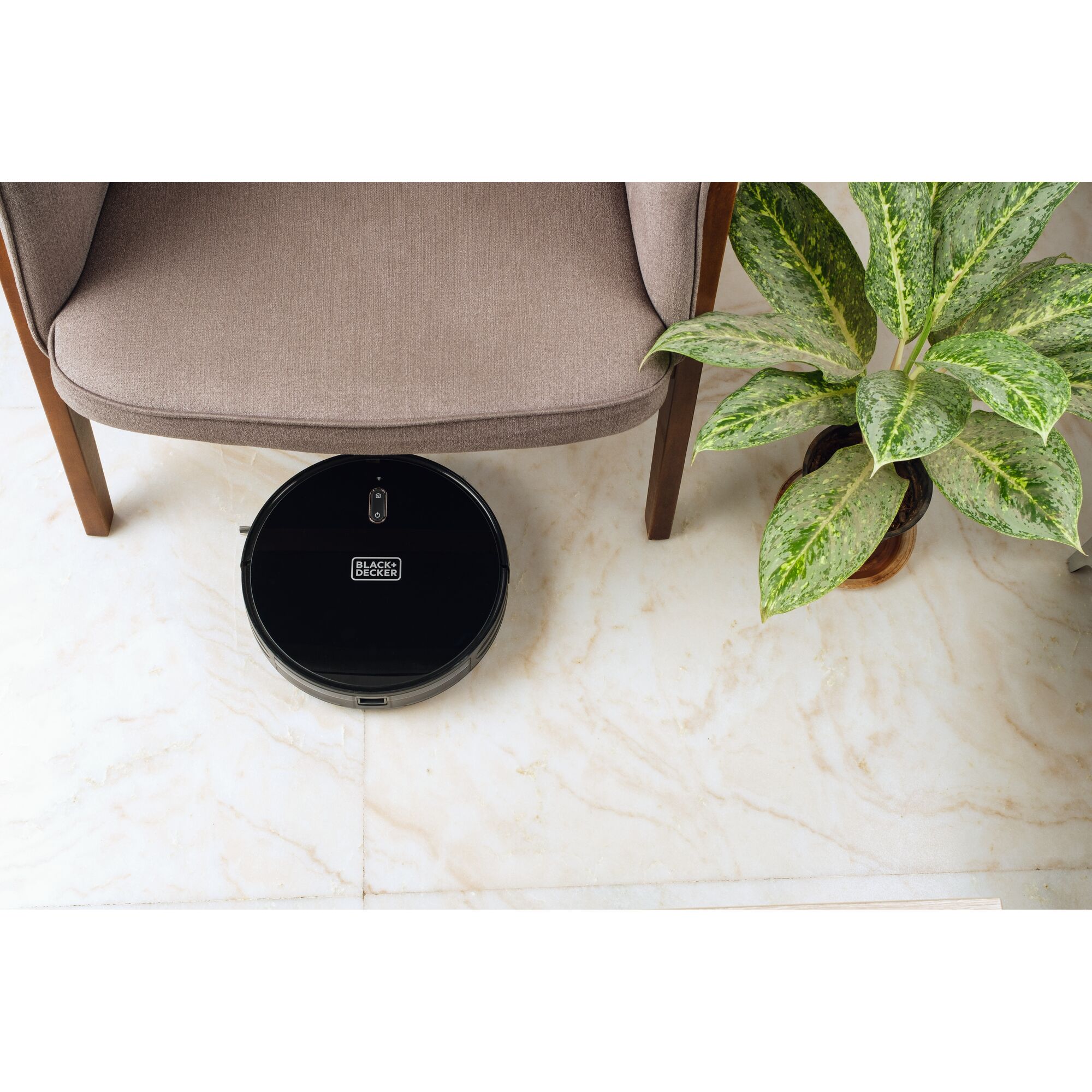 BLACK+DECKER BRVA425B00 robotic vacuum under a brown chair next to a green potted plant on marble floor.