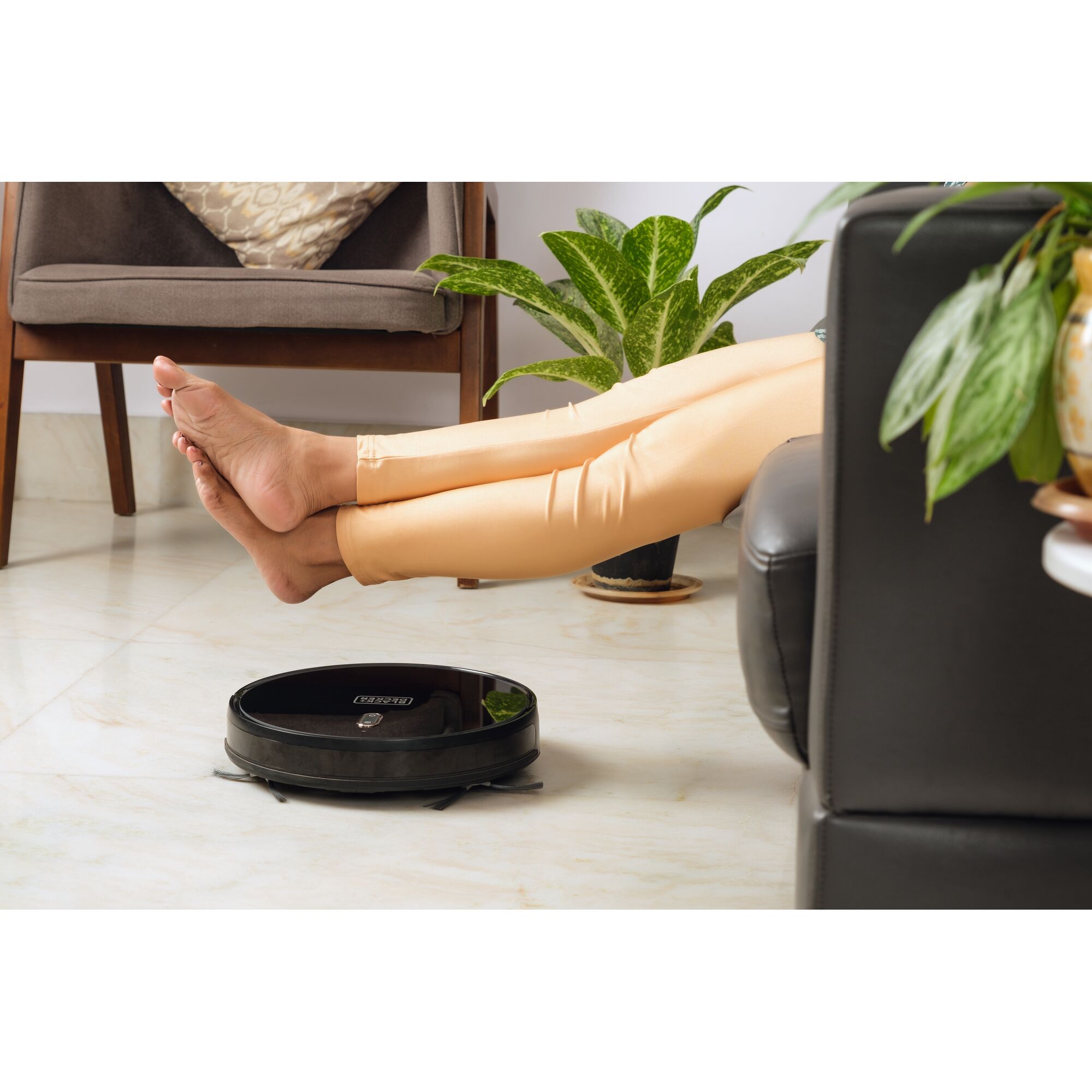 BLACKDECKER BRVA425B00 robot vacuum on floor near legs resting on sofa, plants in background.