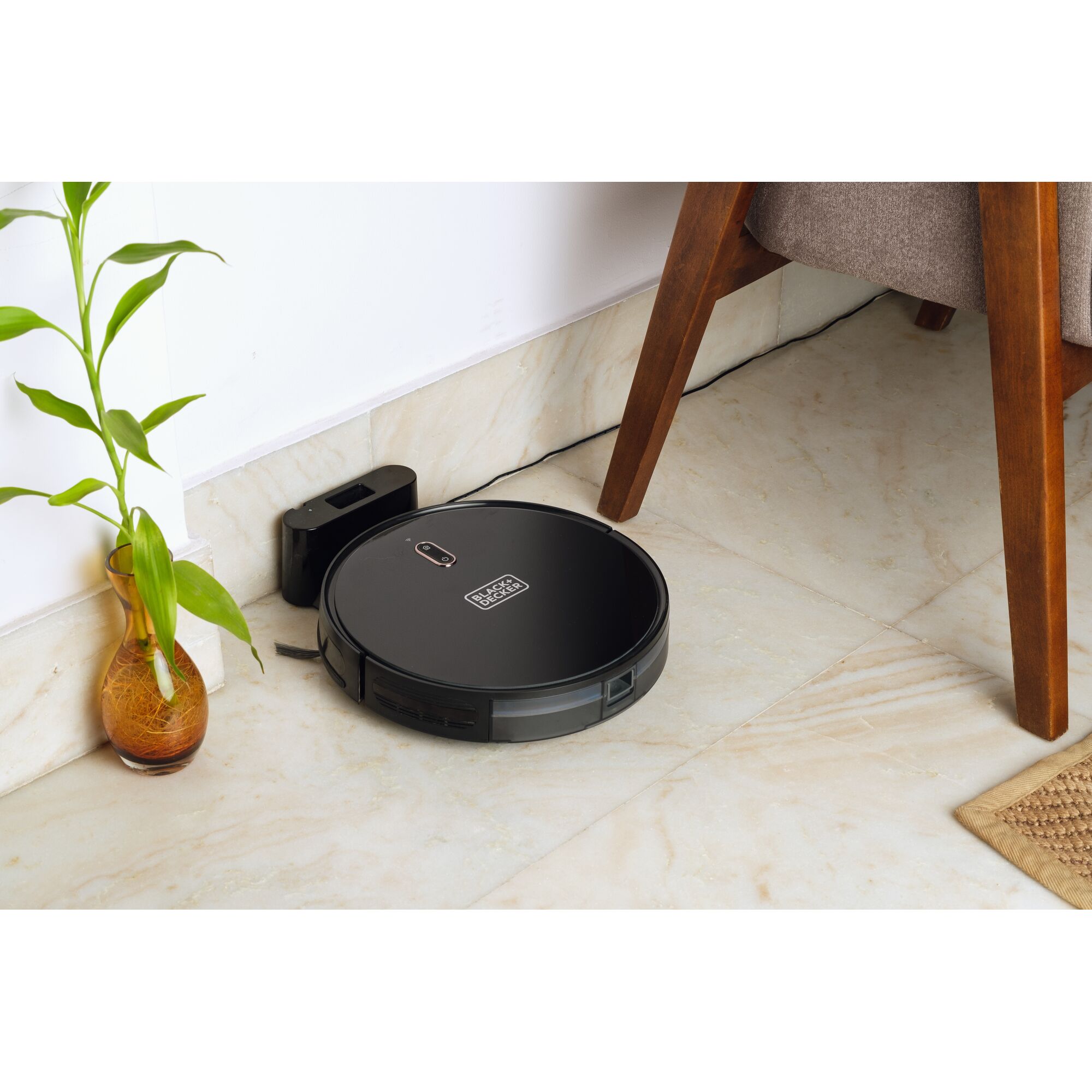 BLACKDECKER BRVA425B00 robotic vacuum in black on tile floor near a wall and chair, with charging dock.