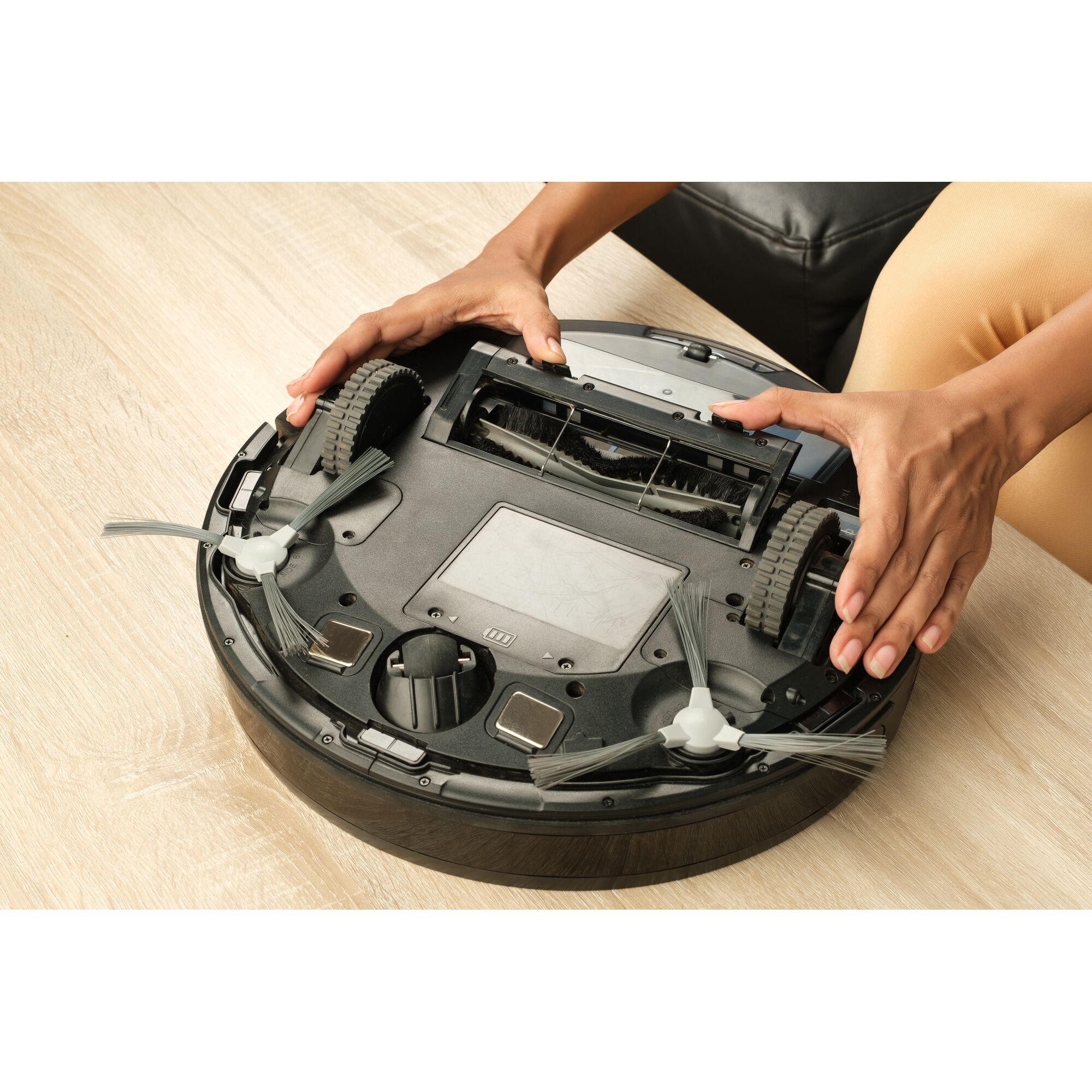 BLACKDECKER BRVA425B00 robot vacuum underside with user hands cleaning side brushes on light wood floor.