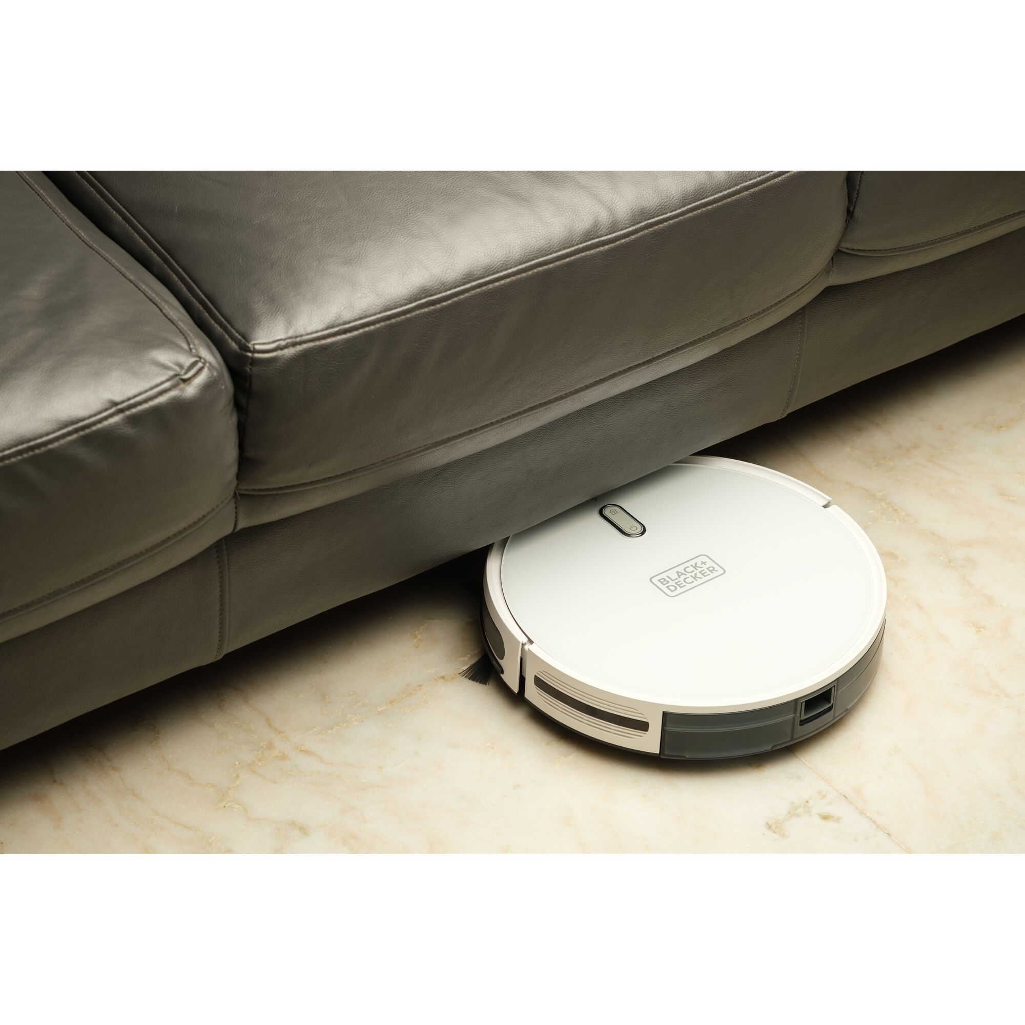 BLACKDECKER BRVA425B10 robot vacuum cleaning under a leather sofa on marble floor, angled view.