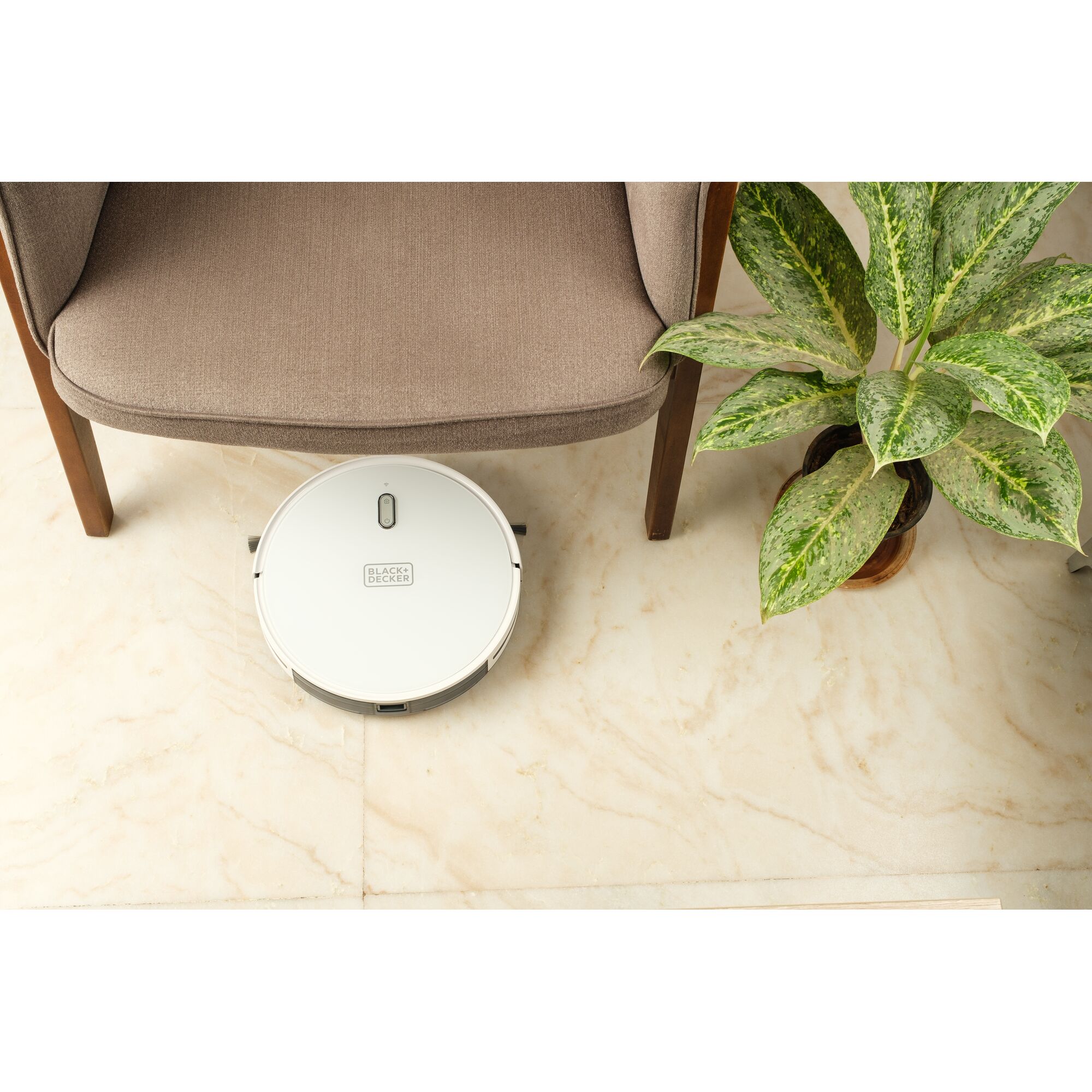 BLACKDECKER BRVA425B10 robot vacuum on marble floor near beige chair and green potted plant, top view.