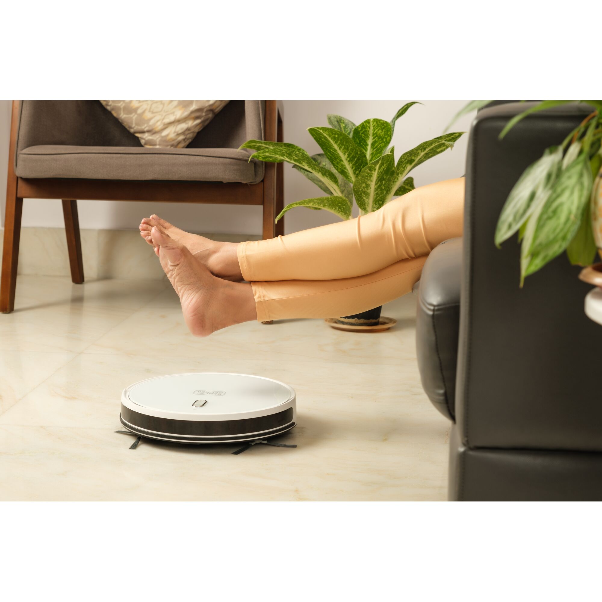 BLACKDECKER BRVA425B10 robot vacuum on floor near seated person’s feet, indoor setting, plants and chair visible.