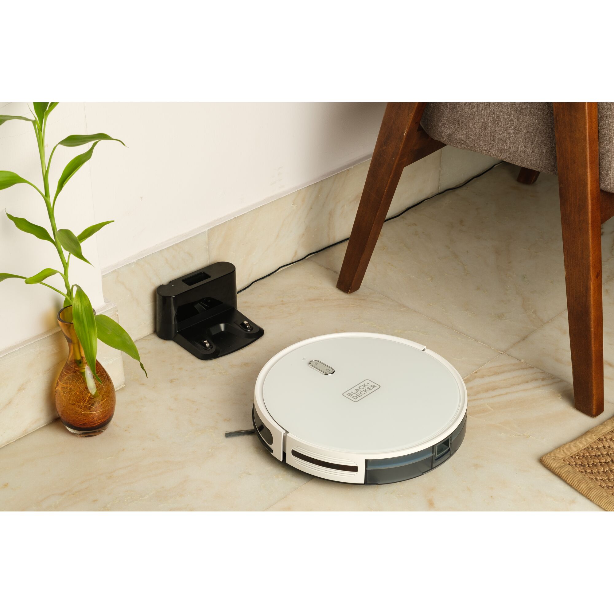 BLACKDECKER BRVA425B10 robotic vacuum near charging dock on marble floor beside chair and plant vase.