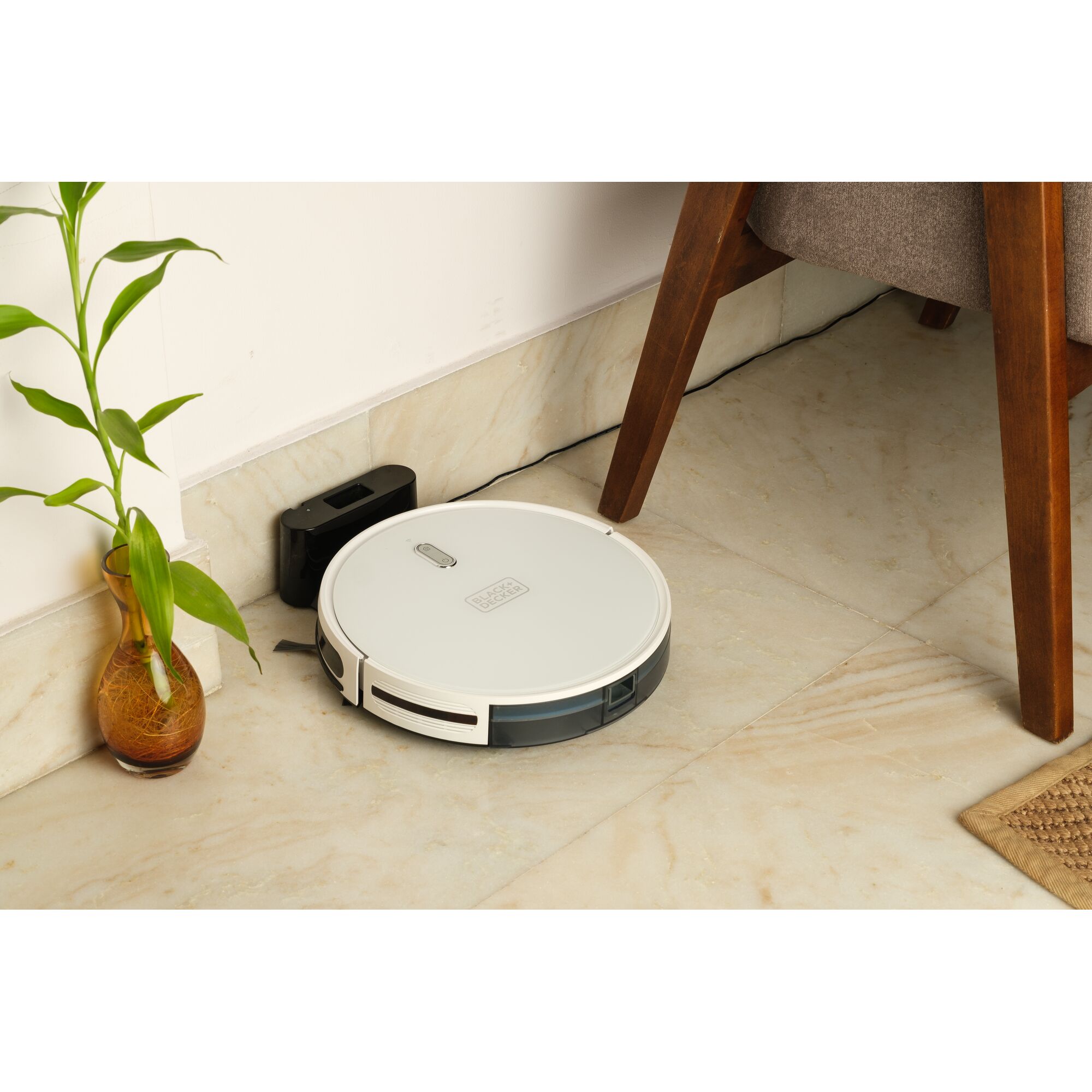 Photo of BLACKDECKER BRVA425B10 robotic vacuum cleaner docking in a corner on tile floor near a plant and wooden furniture.