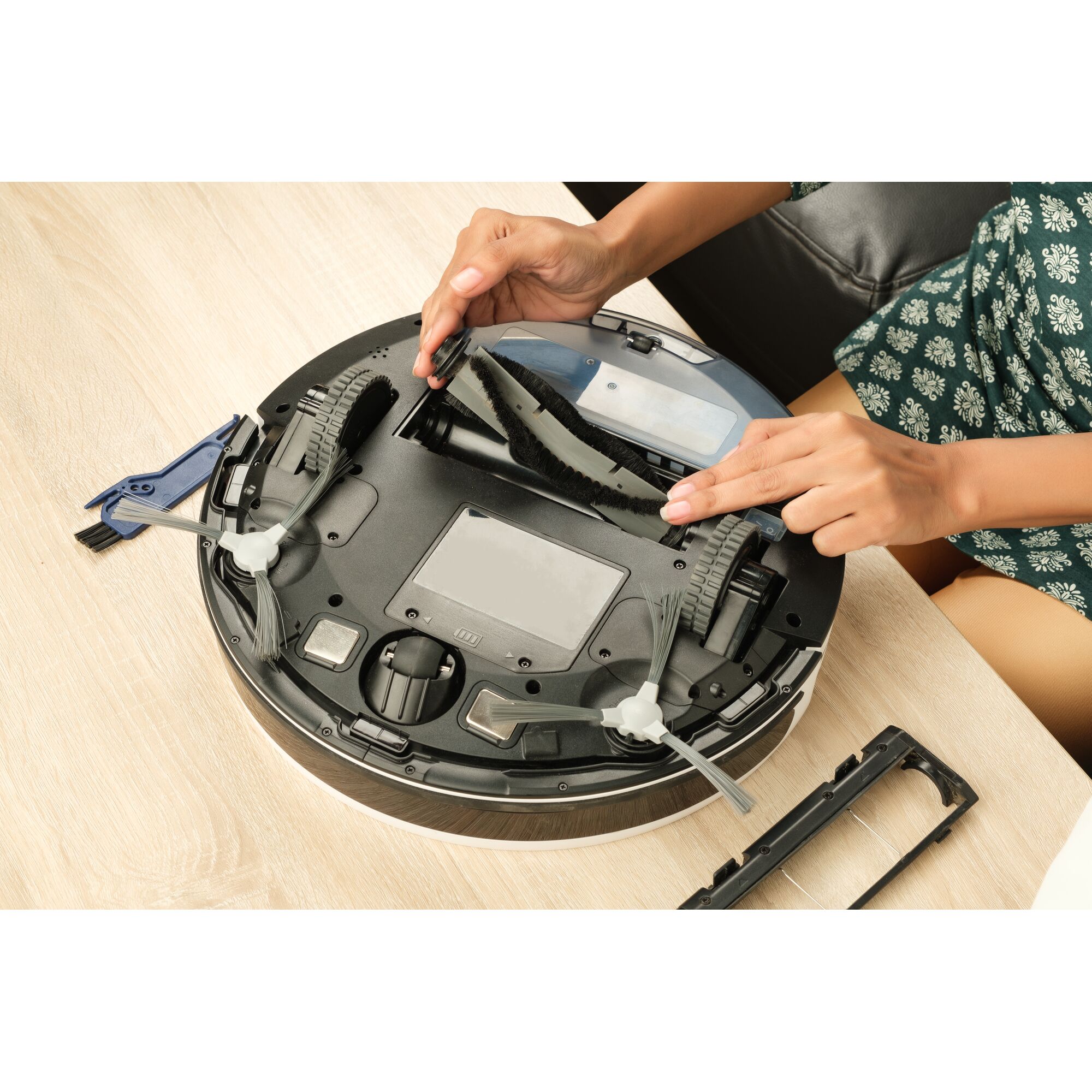 BLACKDECKER BRVA425B10 robot vacuum cleaner bottom view, person cleaning main brush with tool on wooden table.