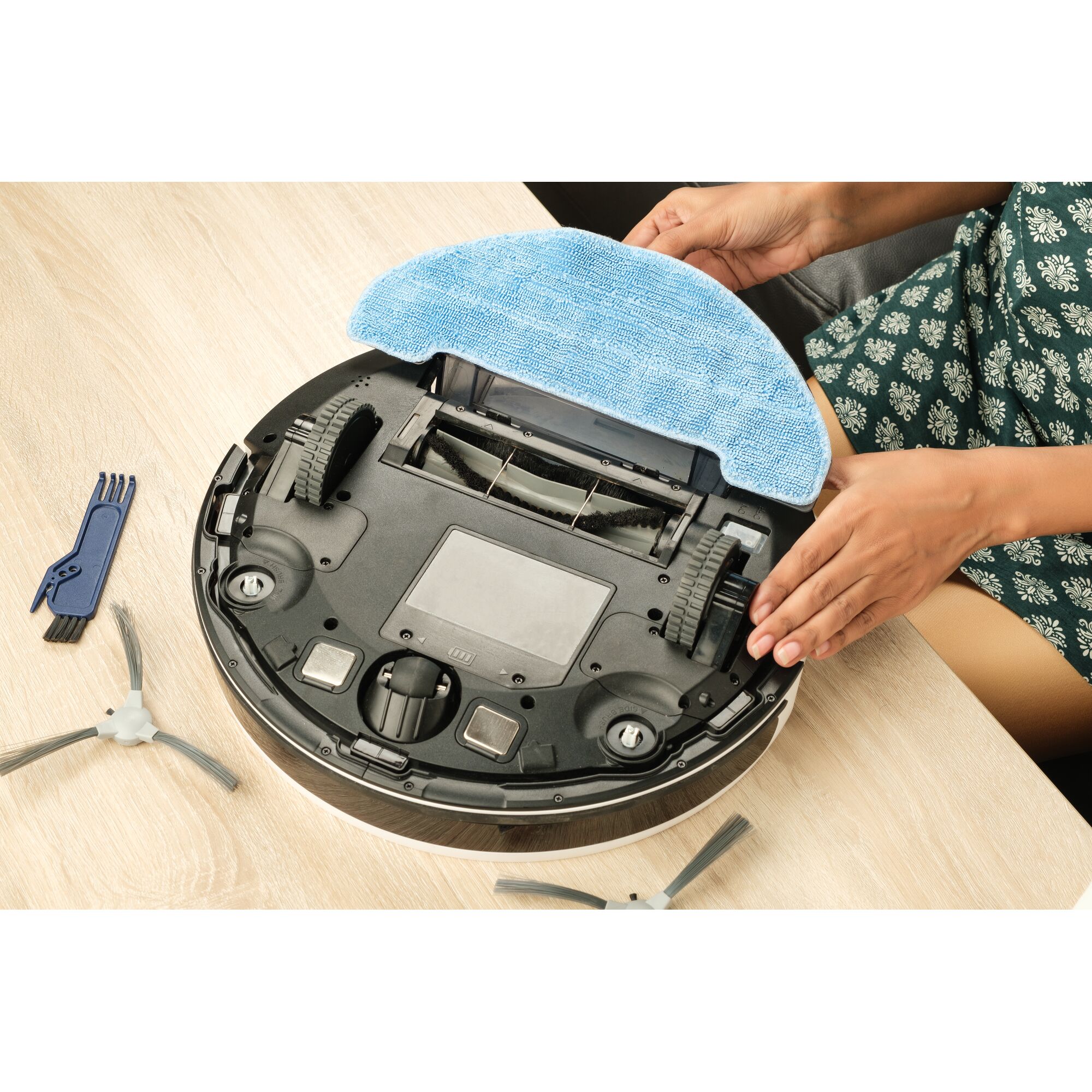 BLACKDECKER BRVA425B10 robot vacuum upside down with blue cleaning pad, extra brushes, and tool on wooden table.