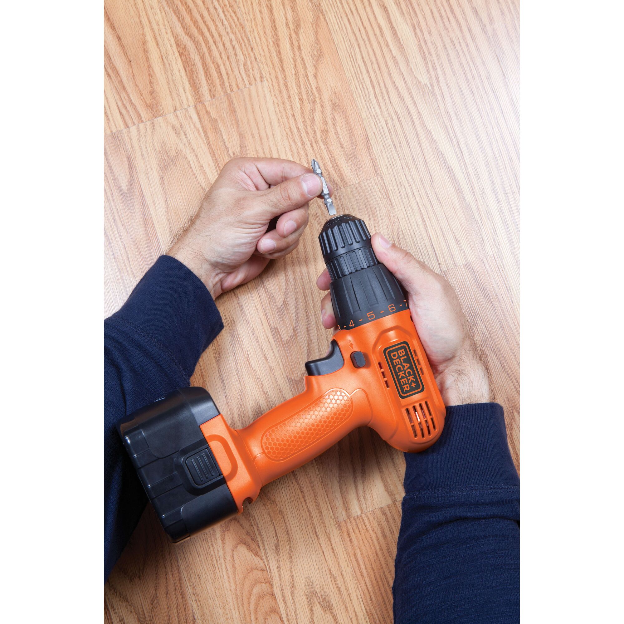BLACKDECKER CD121K50 cordless drill held by hands on wood floor, orange and black design, side view.