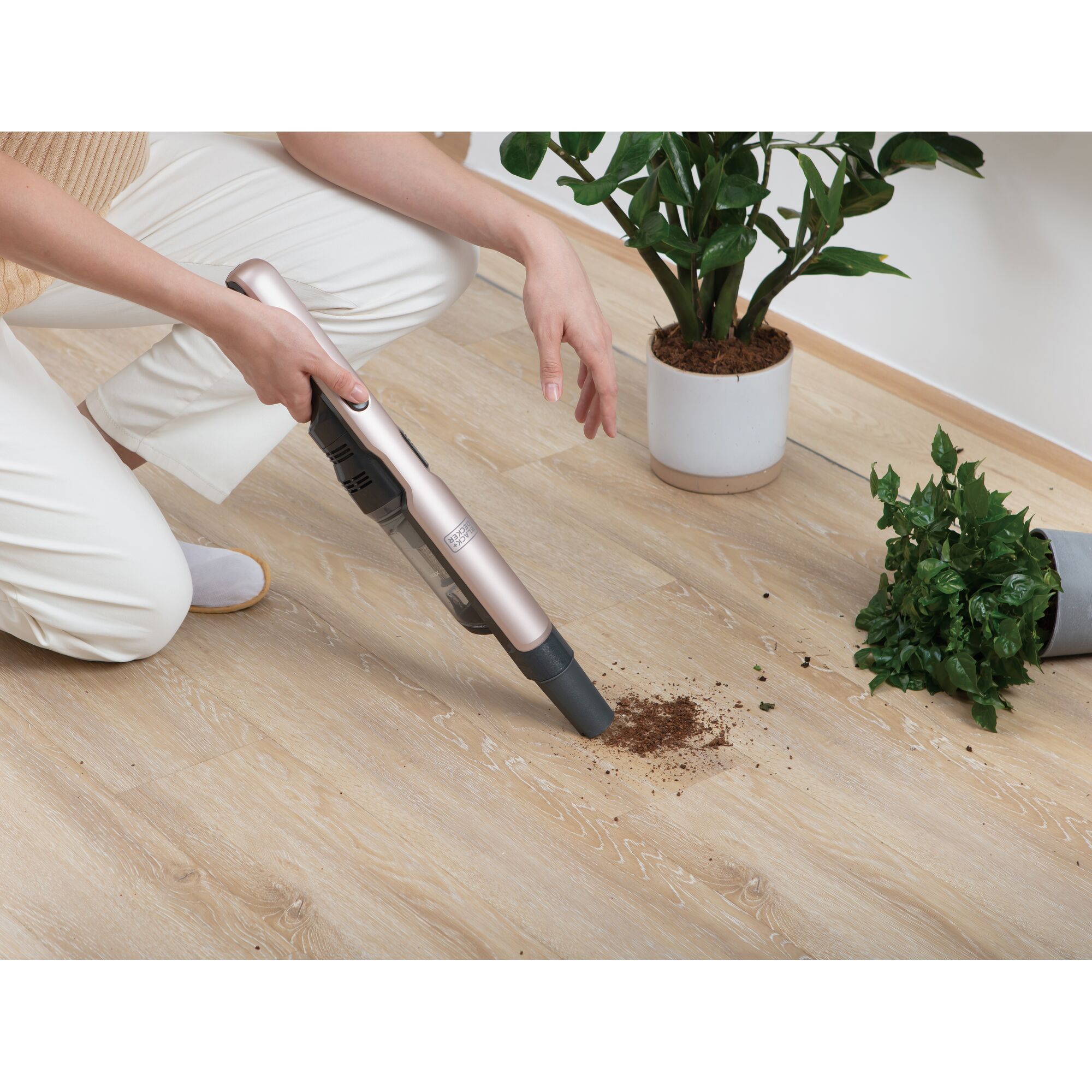 BLACKDECKER DVC320B24 handheld vacuum cleaner cleaning spilled soil near plants on wooden floor, angled view.