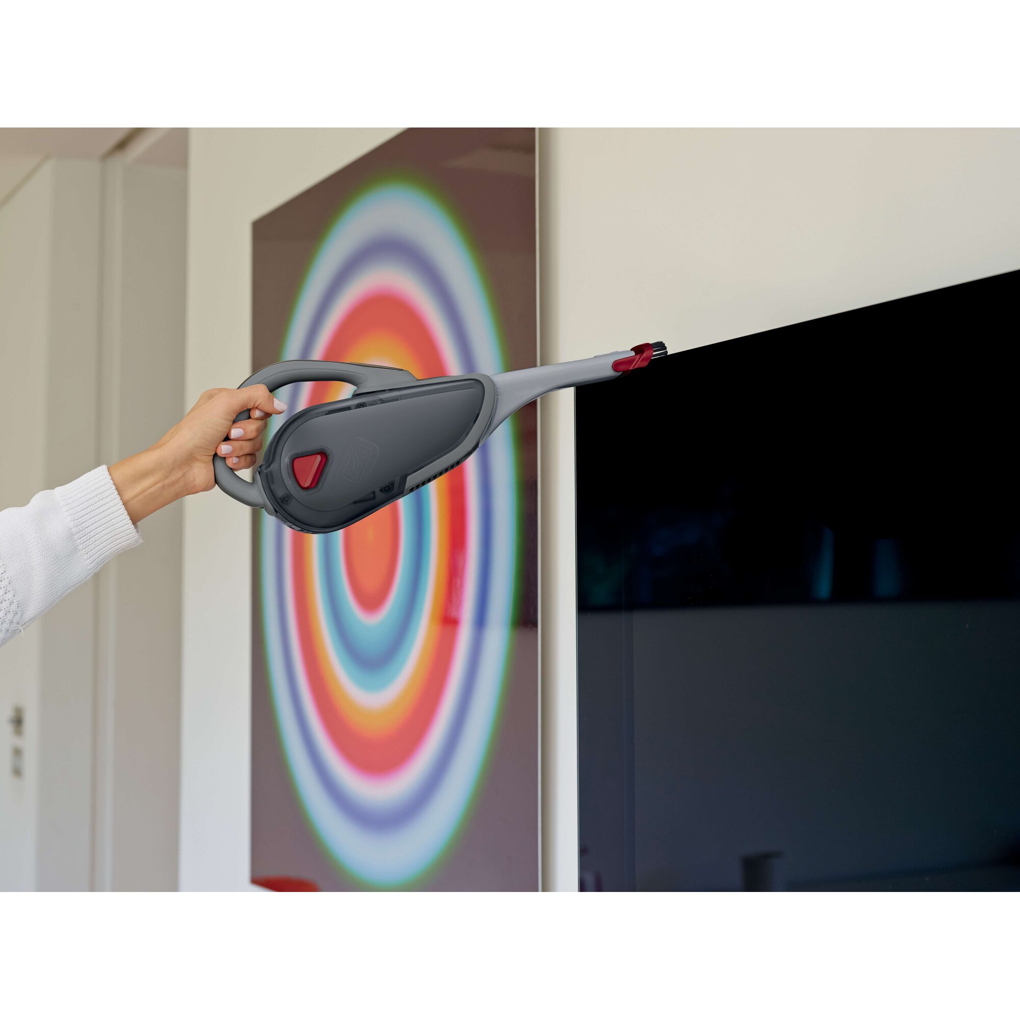 A person using a BLACK+DECKER handheld vacuum cleaner with an extended nozzle attachment to clean around a television screen, with a colorful concentric circle artwork in the background.