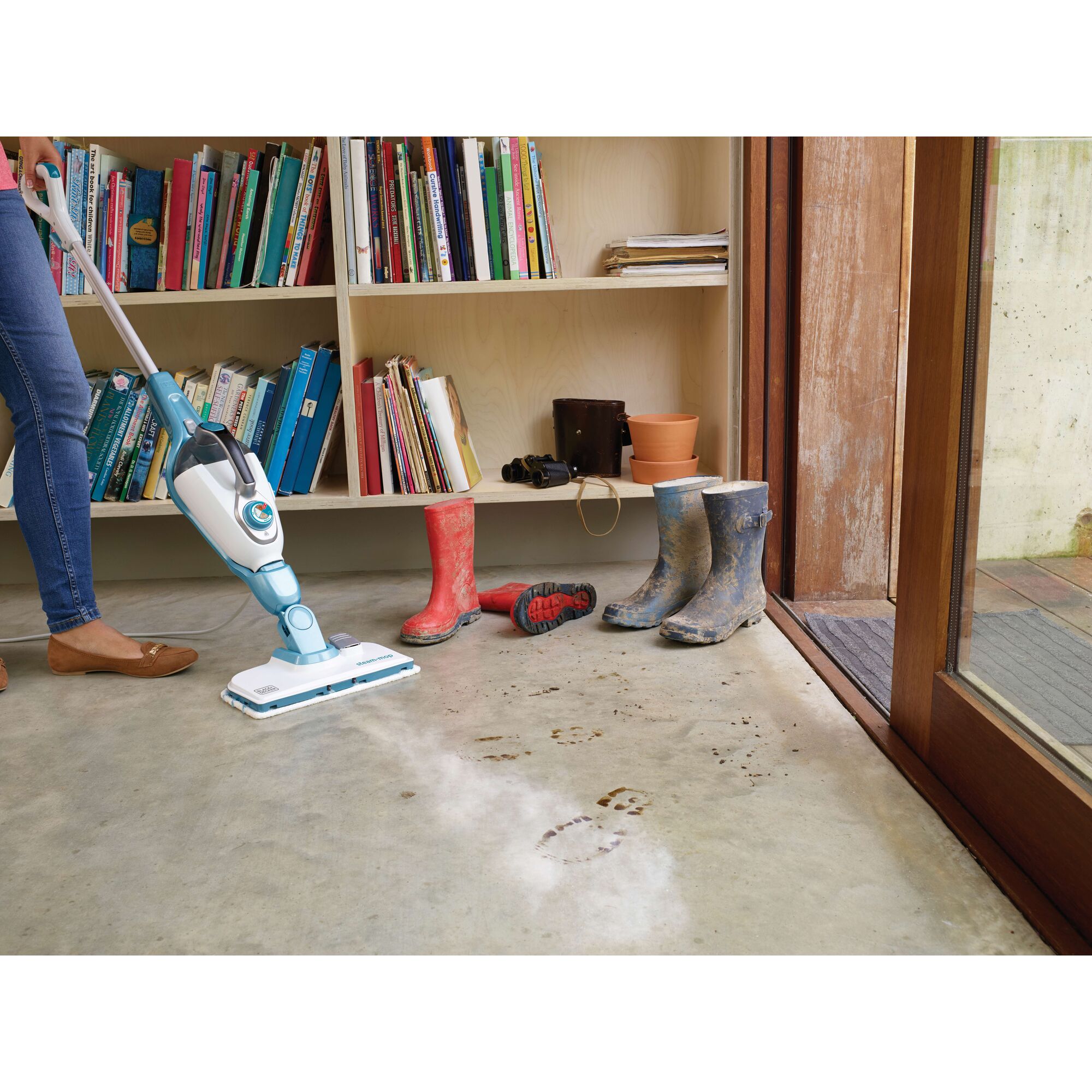 A person uses a BLACK+DECKER steam mop to clean muddy footprints off a concrete floor near a bookshelf and pairs of dirty boots.