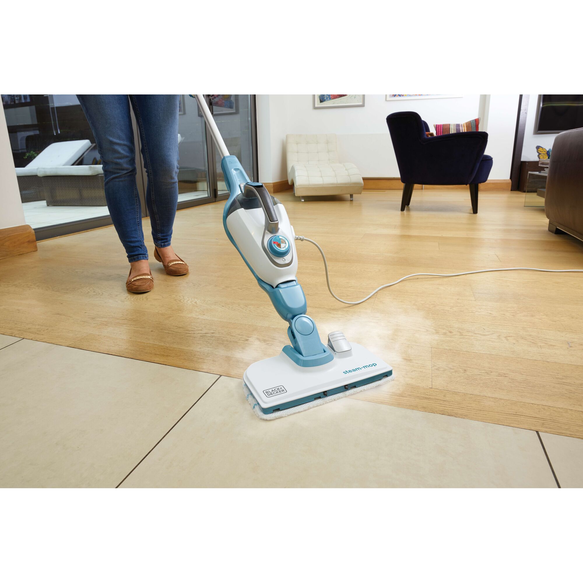 A person using a BLACK+DECKER steam mop to clean a tiled and wooden floor in a modern living room.