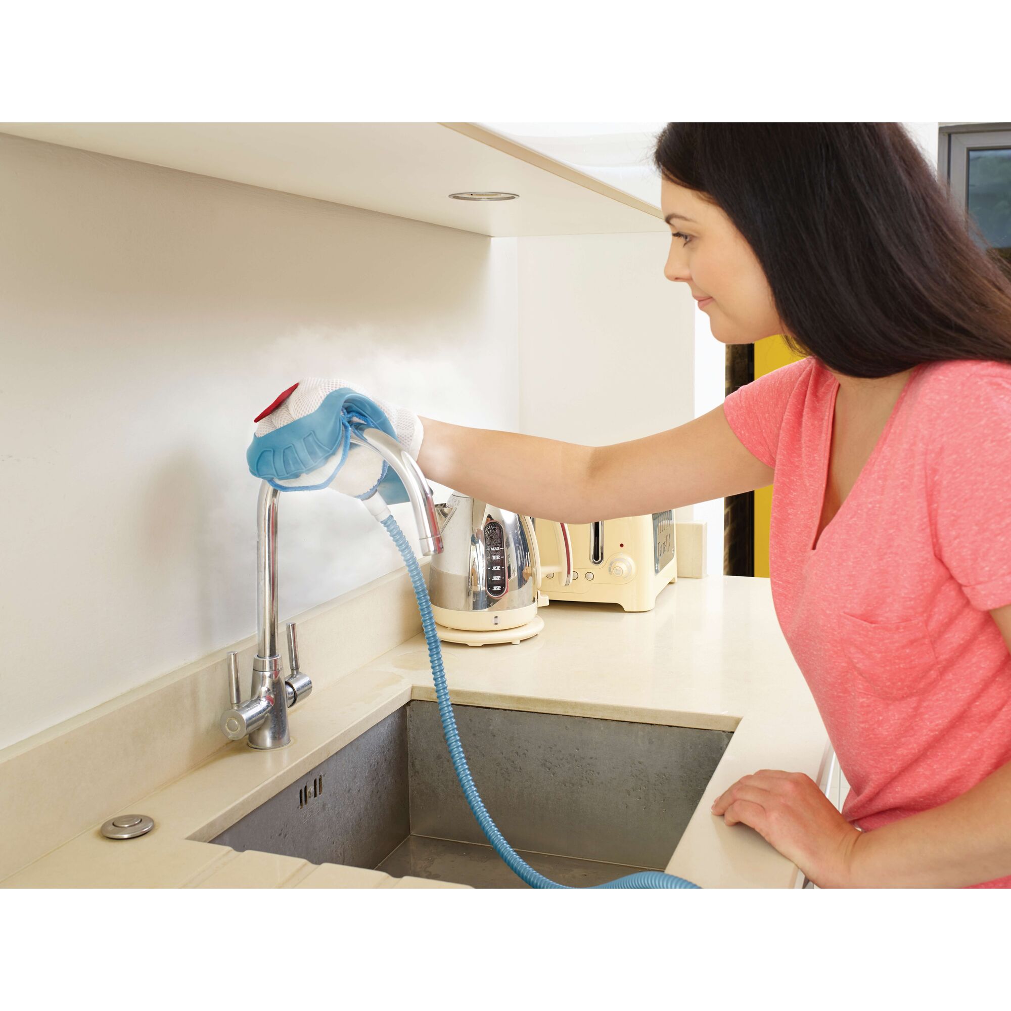 Person in a pink shirt using a steam cleaning glove to clean a kitchen faucet and counter. The steam cleaning glove is connected to a blue hose. The person's face is blurred for privacy.