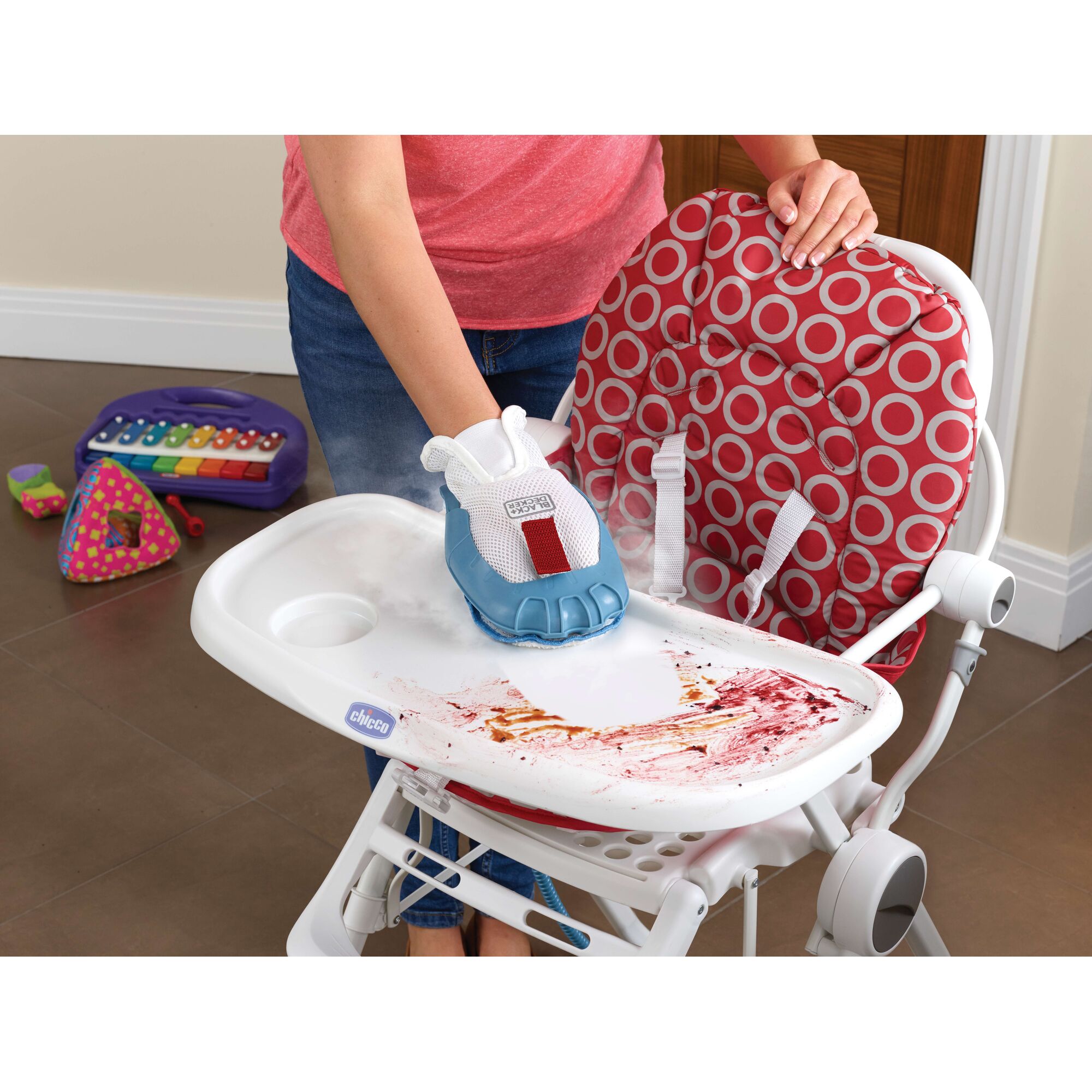 A person cleans a dirty high chair tray with a steam cleaning glove from BLACK+DECKER. The tray has food stains, and children's toys are visible on the floor in the background.