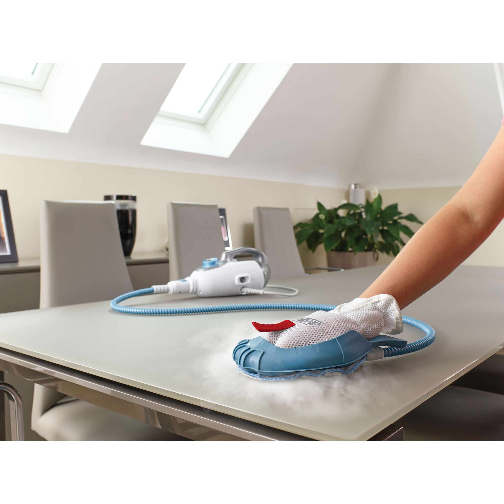 A person cleans a tabletop in a modern room using a BLACK+DECKER steam cleaning device with a blue hand attachment, releasing visible steam.
