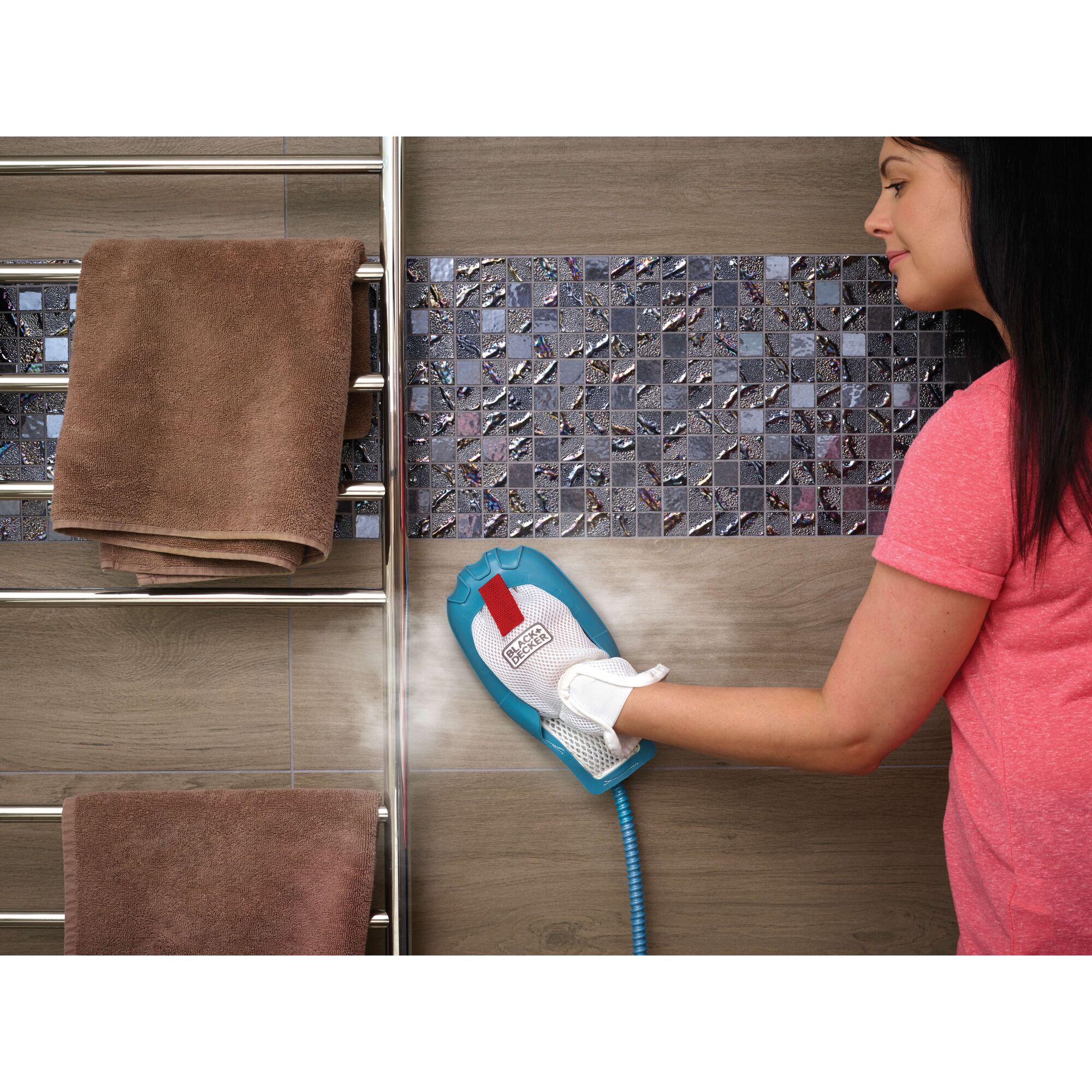 A person uses a BLACK+DECKER steam cleaning glove on tile wall in a bathroom. There are brown towels hanging on a metal rack next to the wall.