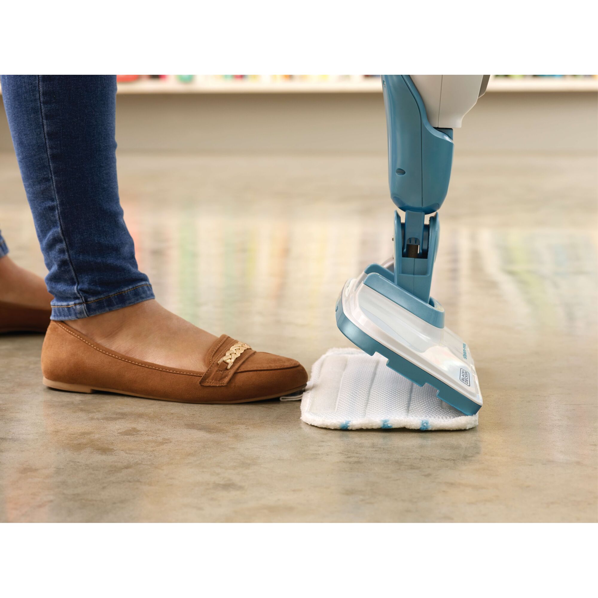 A close-up of a person wearing brown shoes and jeans using their foot to attach a cleaning pad to a blue BLACK+DECKER steam mop on a smooth floor.