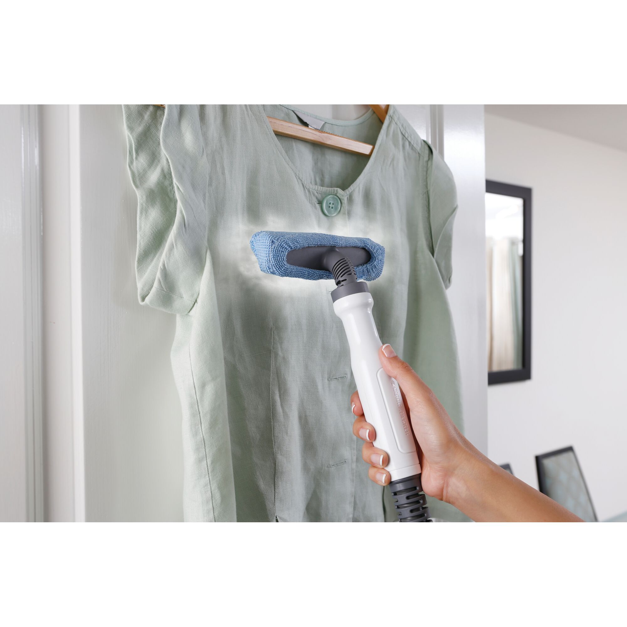 A hand is holding a steam cleaning device with a blue cloth attachment, cleaning or steaming a light green shirt hanging on a hanger in a doorway.
