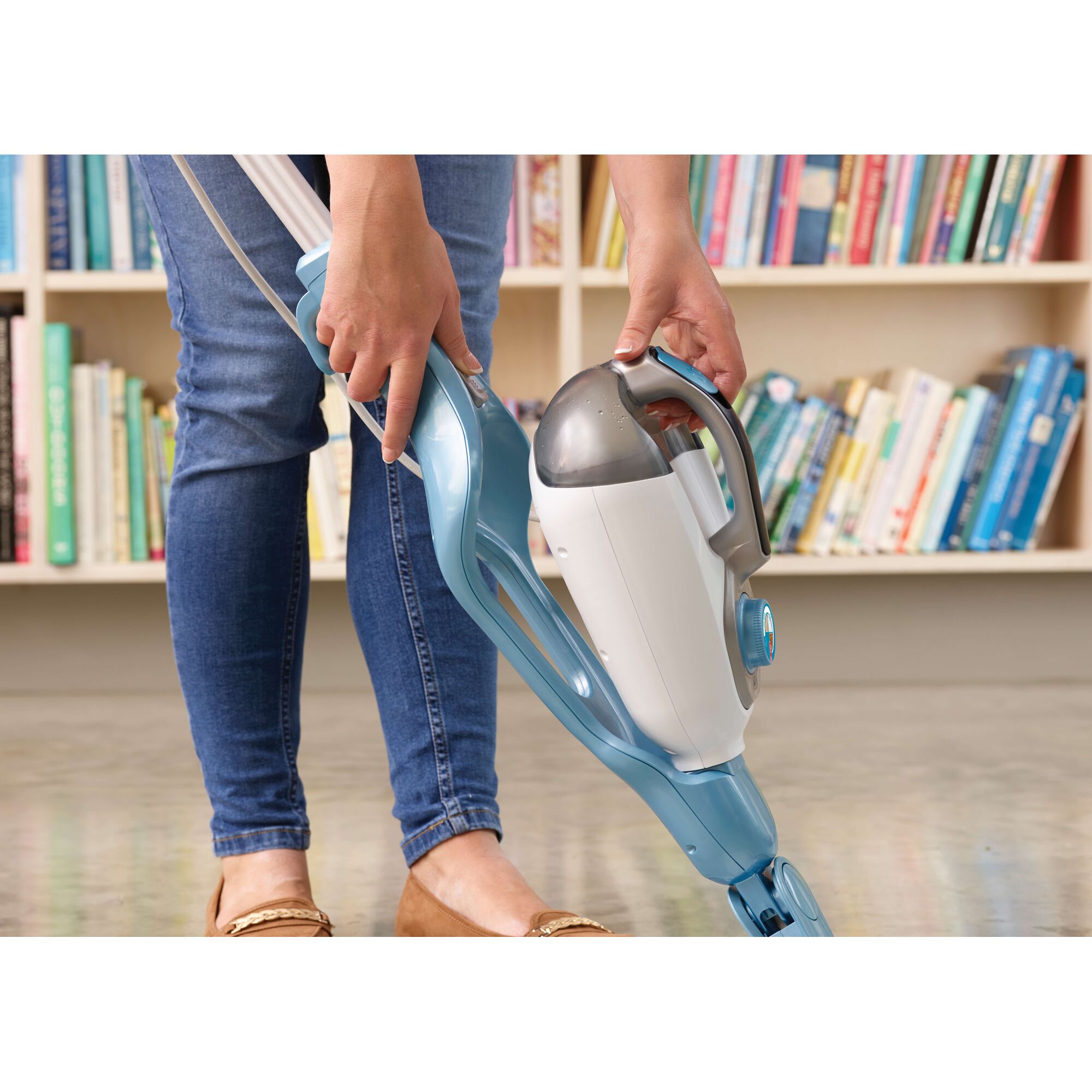 A person using a BLACK+DECKER steam mop with detachable handheld unit, standing on a polished floor in front of a bookshelf filled with books.