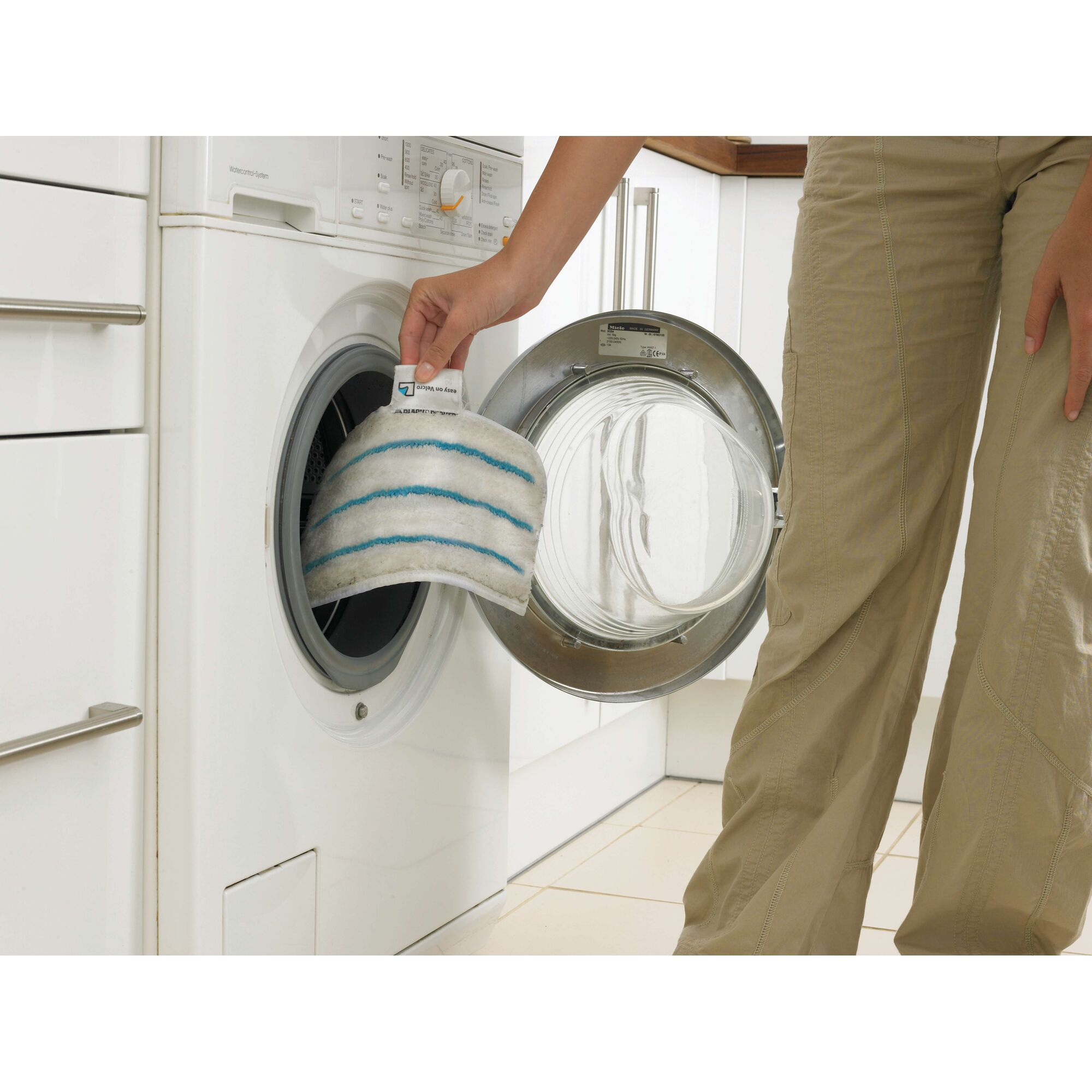 A person is placing a white cleaning pad with blue stripes into the drum of a front-loading washing machine.