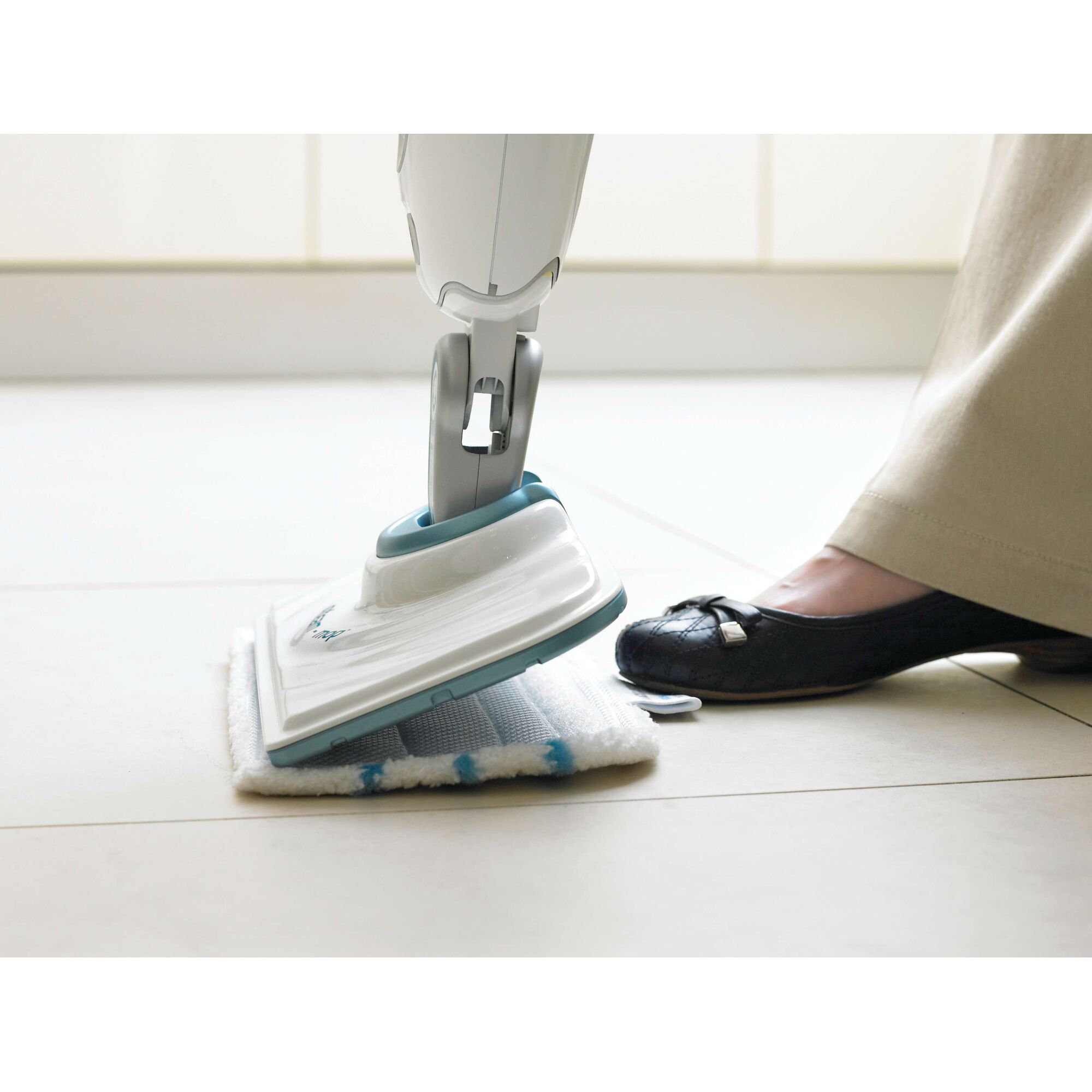 A close-up of a person using a BLACK+DECKER steam mop on a tiled floor. The person's foot is pressing a button on the mop, and the mop head is equipped with a cleaning pad.
