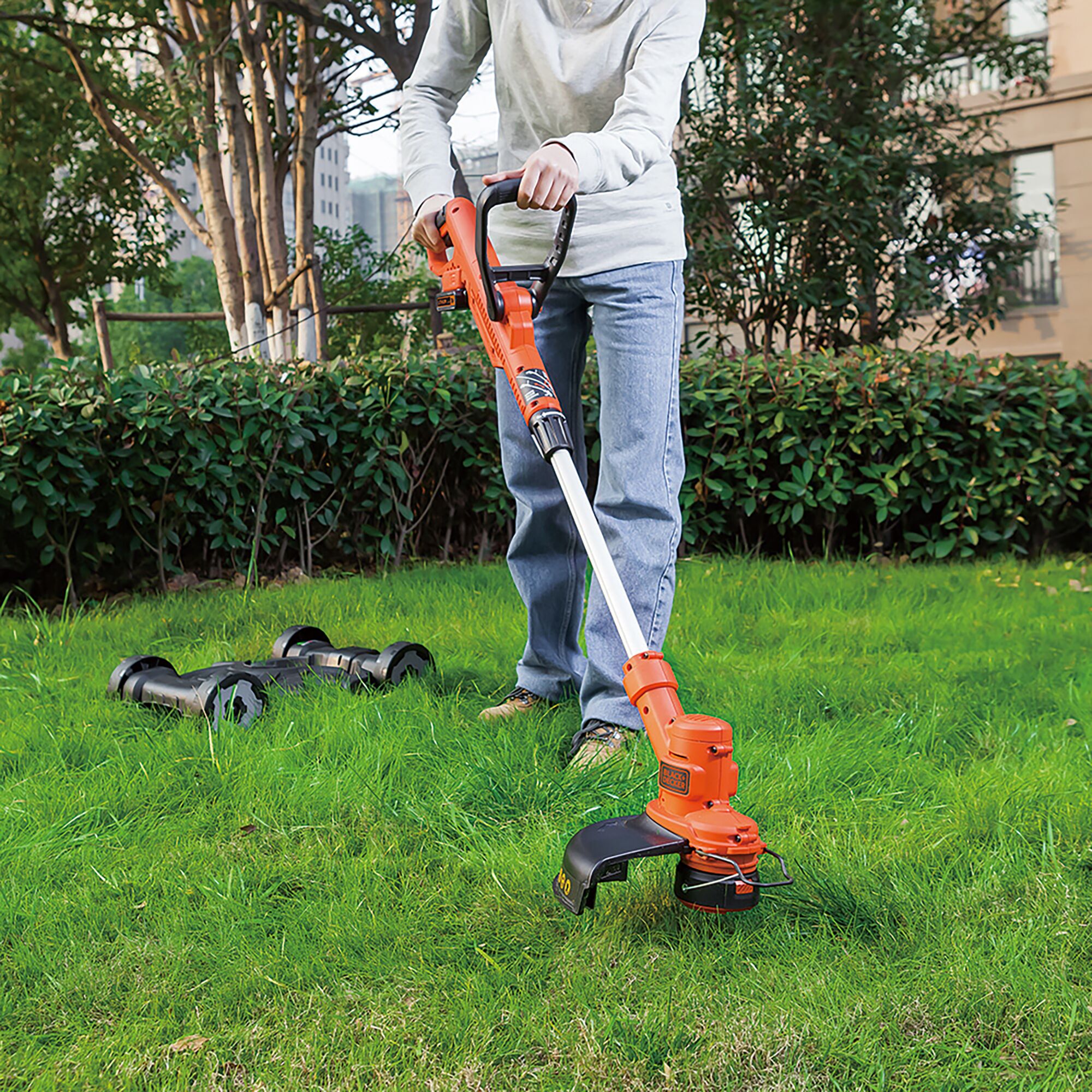 BLACKDECKER GCM18BN cordless grass trimmer in use on lawn, orange and black design, garden background.