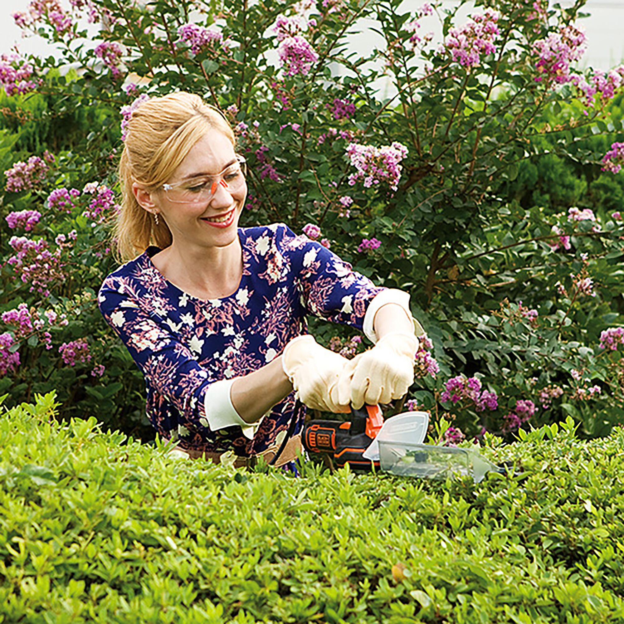 BLACKDECKER GSH1000 hedge trimmer held by person trimming green bushes in garden with flowering plants.