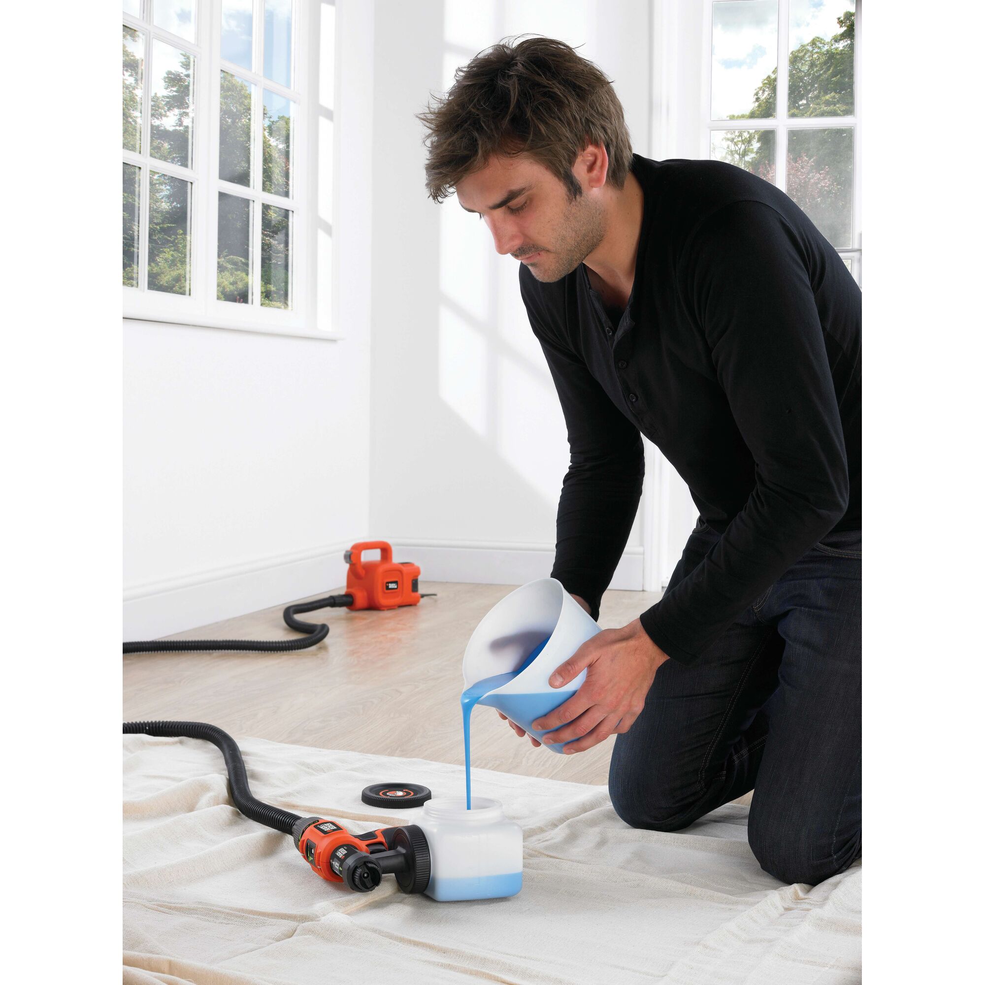 A person kneeling on a sheet indoors, pouring blue liquid into the container of a BLACK+DECKER HVLP400 paint sprayer. An orange paint sprayer unit and hose are visible on the floor, with large windows in the background.